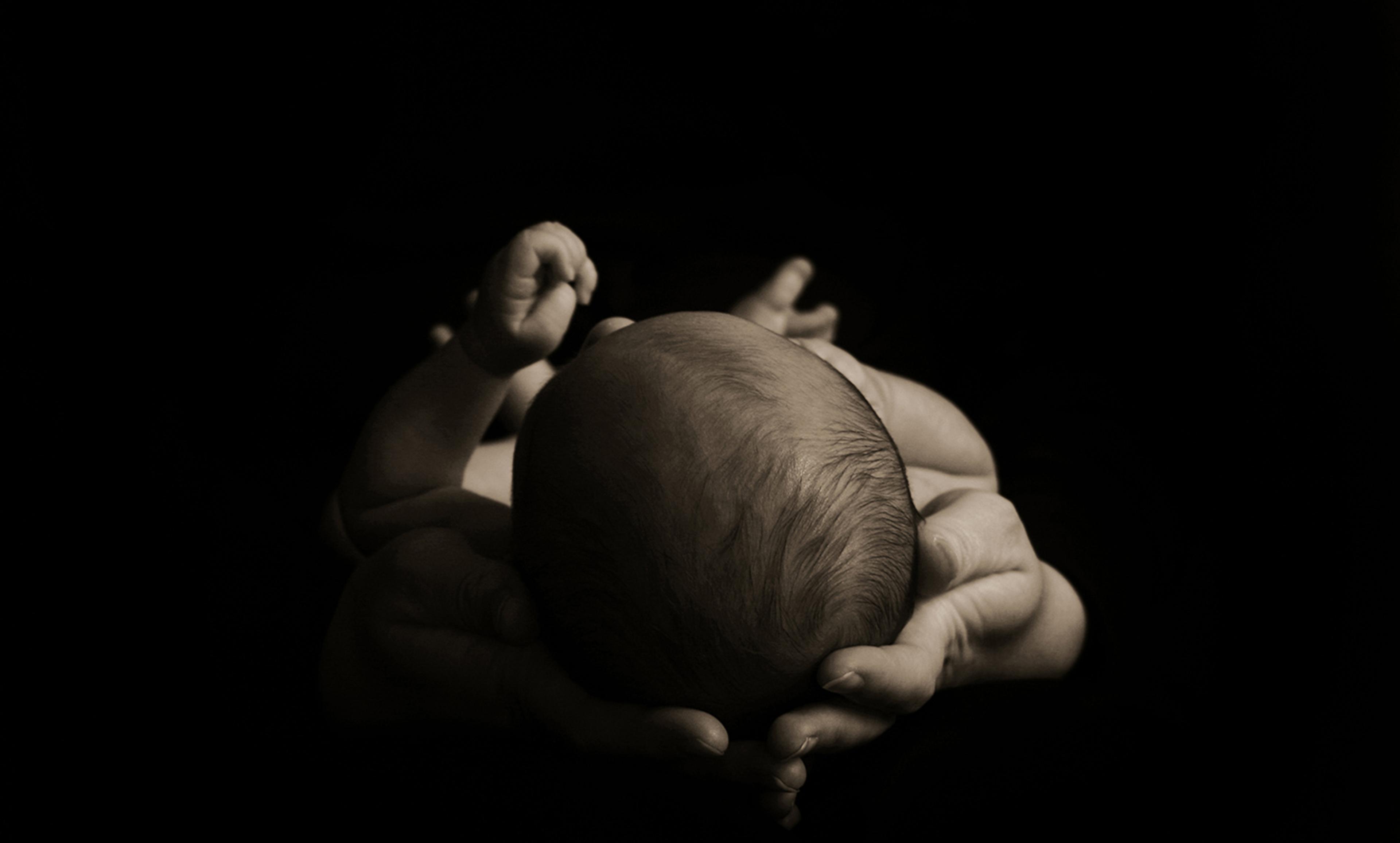 A black-and-white photo of a baby seen from above cradled in hands in low light against a dark background.