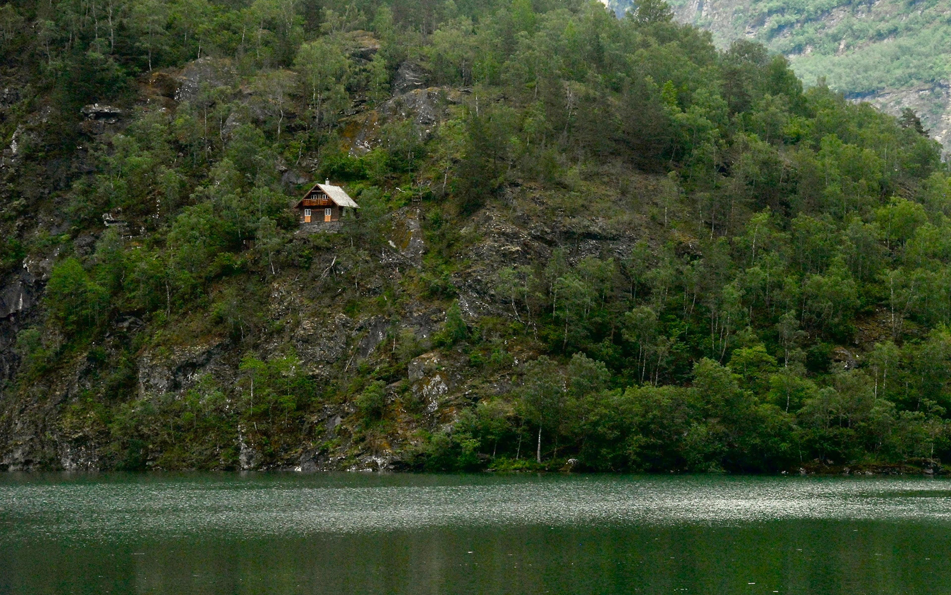 A wooden cabin perched on a forested hillside above a tranquil lake, surrounded by dense trees and rugged terrain.
