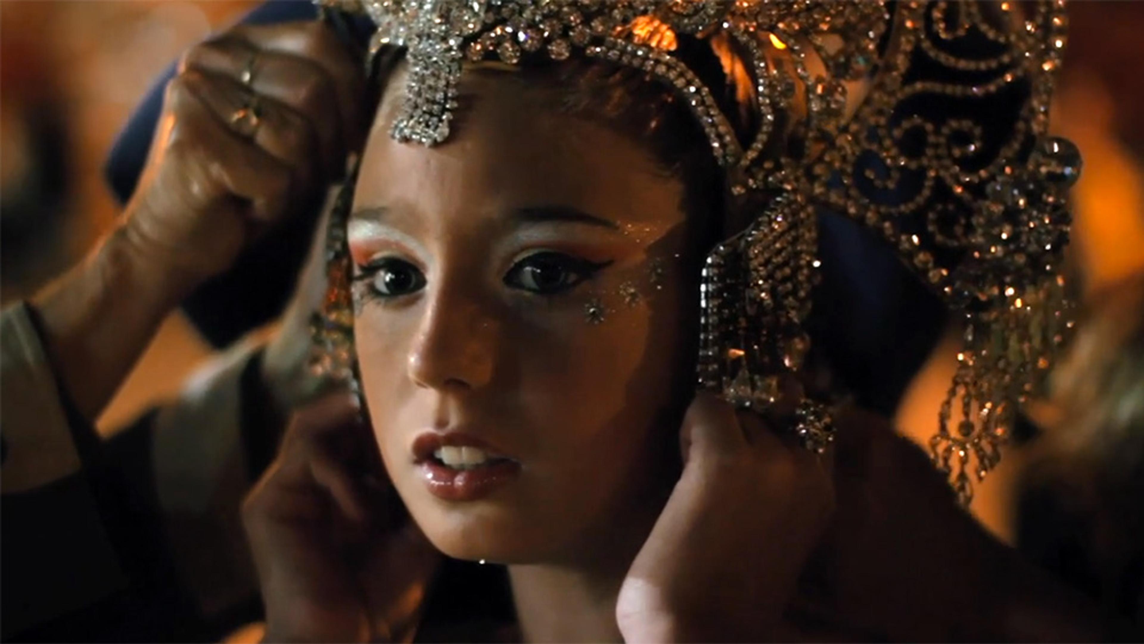 A photo of a young girl having an ornate headpiece adjusted, with a close-up on her face, showing detailed makeup and decorations.