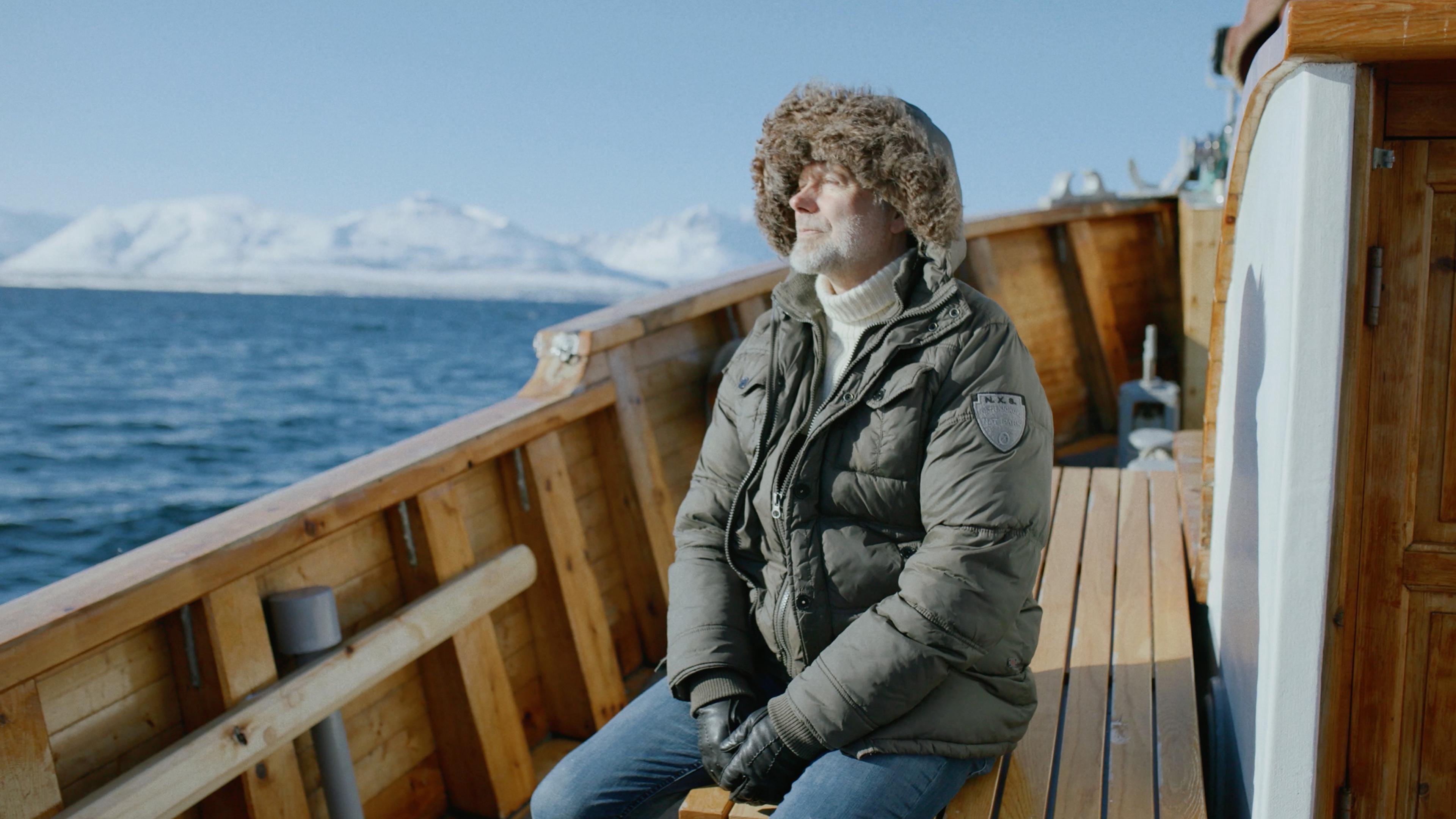 A man in winter clothing sits on a wooden boat, eyes half closed, with a snowy mountain landscape and water in the background.