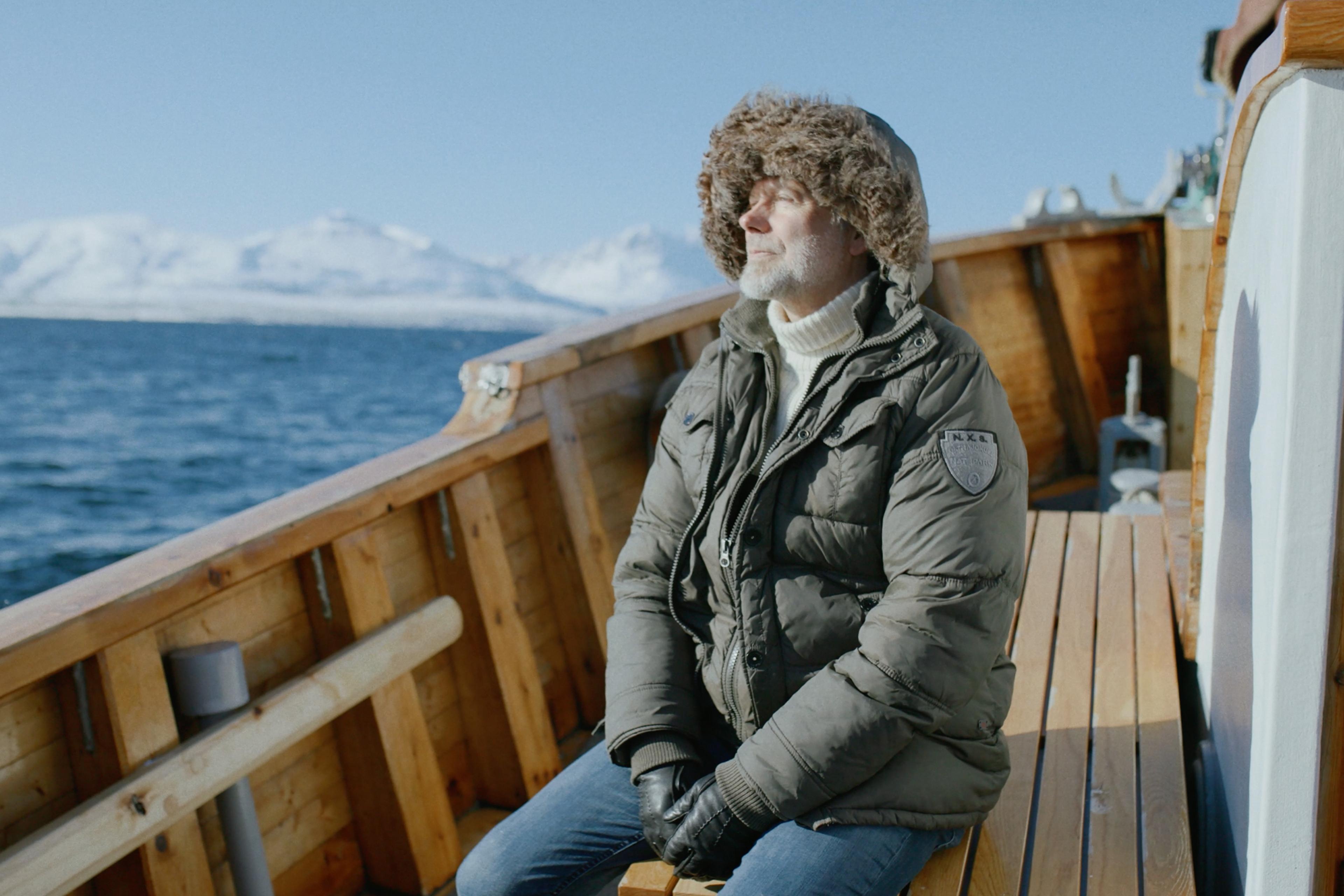 A man in winter clothing sits on a wooden boat, eyes half closed, with a snowy mountain landscape and water in the background.