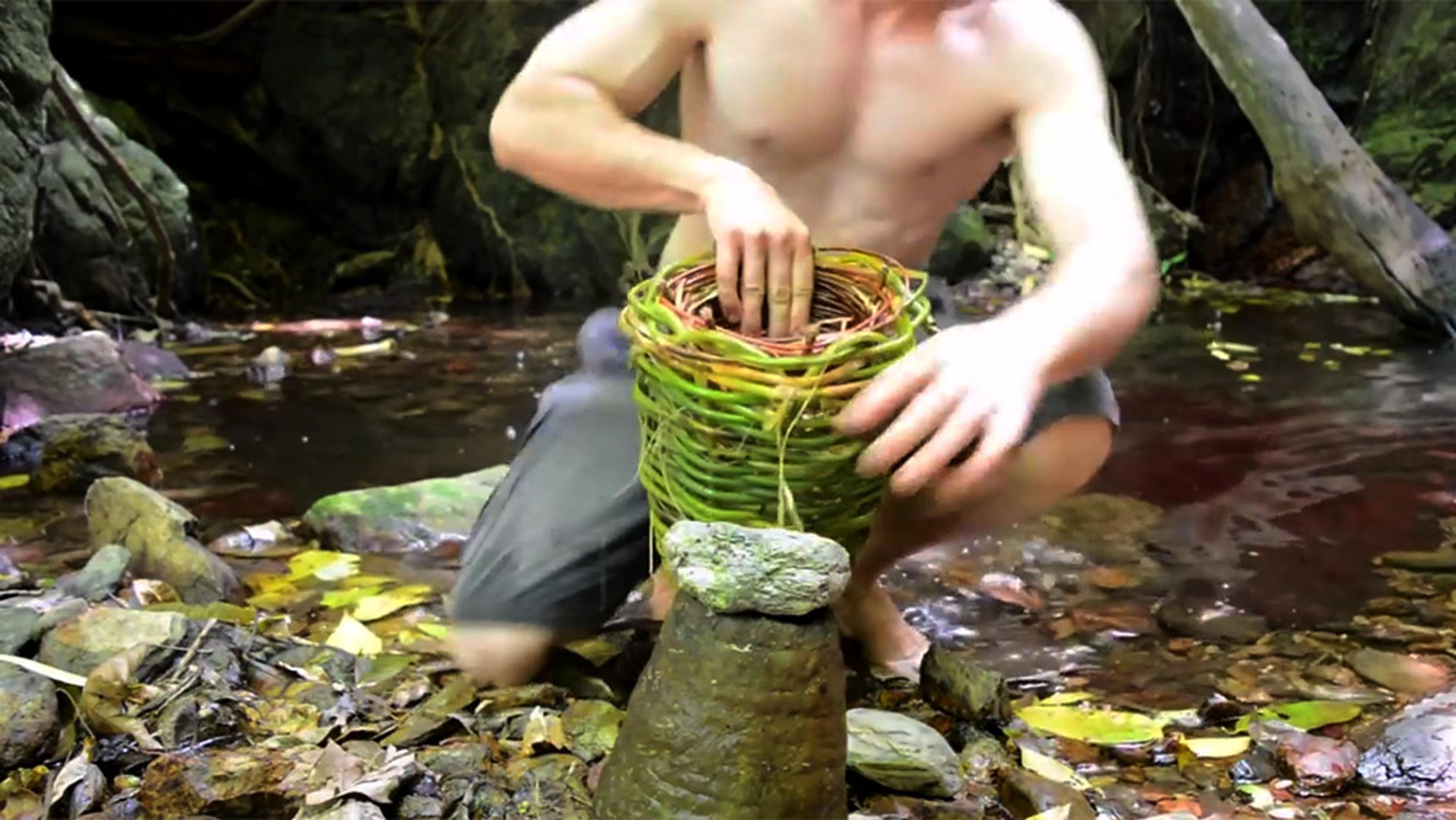 A shirtless man kneels by a stream weaving a green basket. The scene is set in a forest with rocks and leaves around.