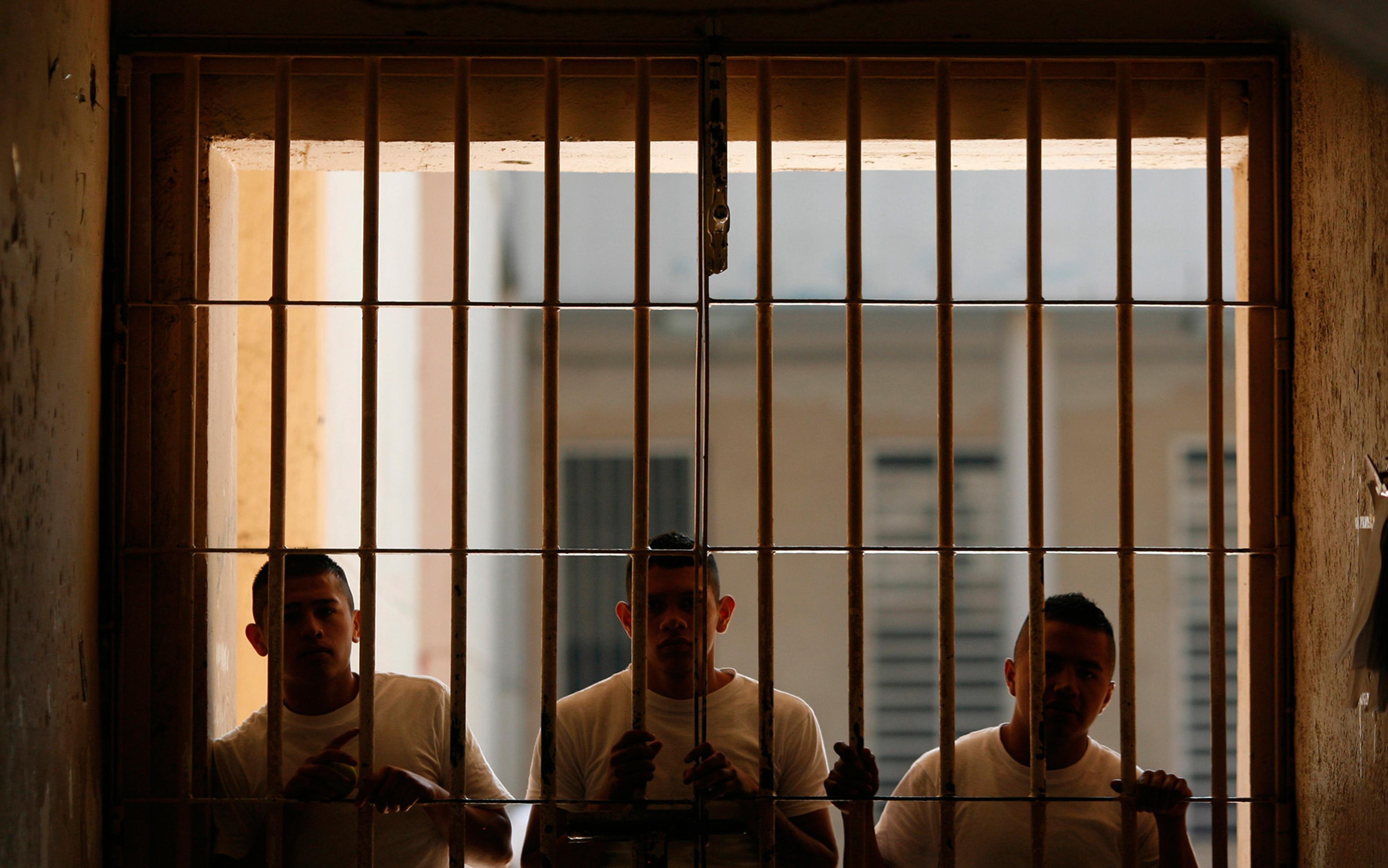 Three men in white T-shirts standing behind prison bars in a dimly lit space.