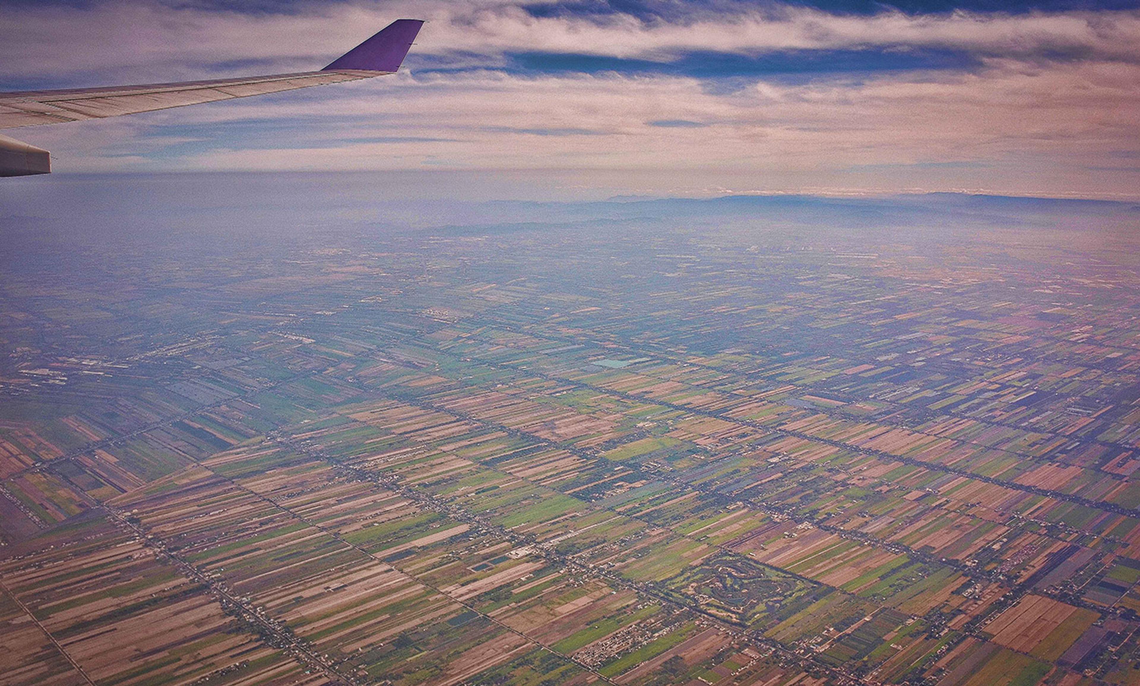 Aerial photo of patchwork farmland taken from an aeroplane window, with a portion of the wing visible against a cloudy sky.