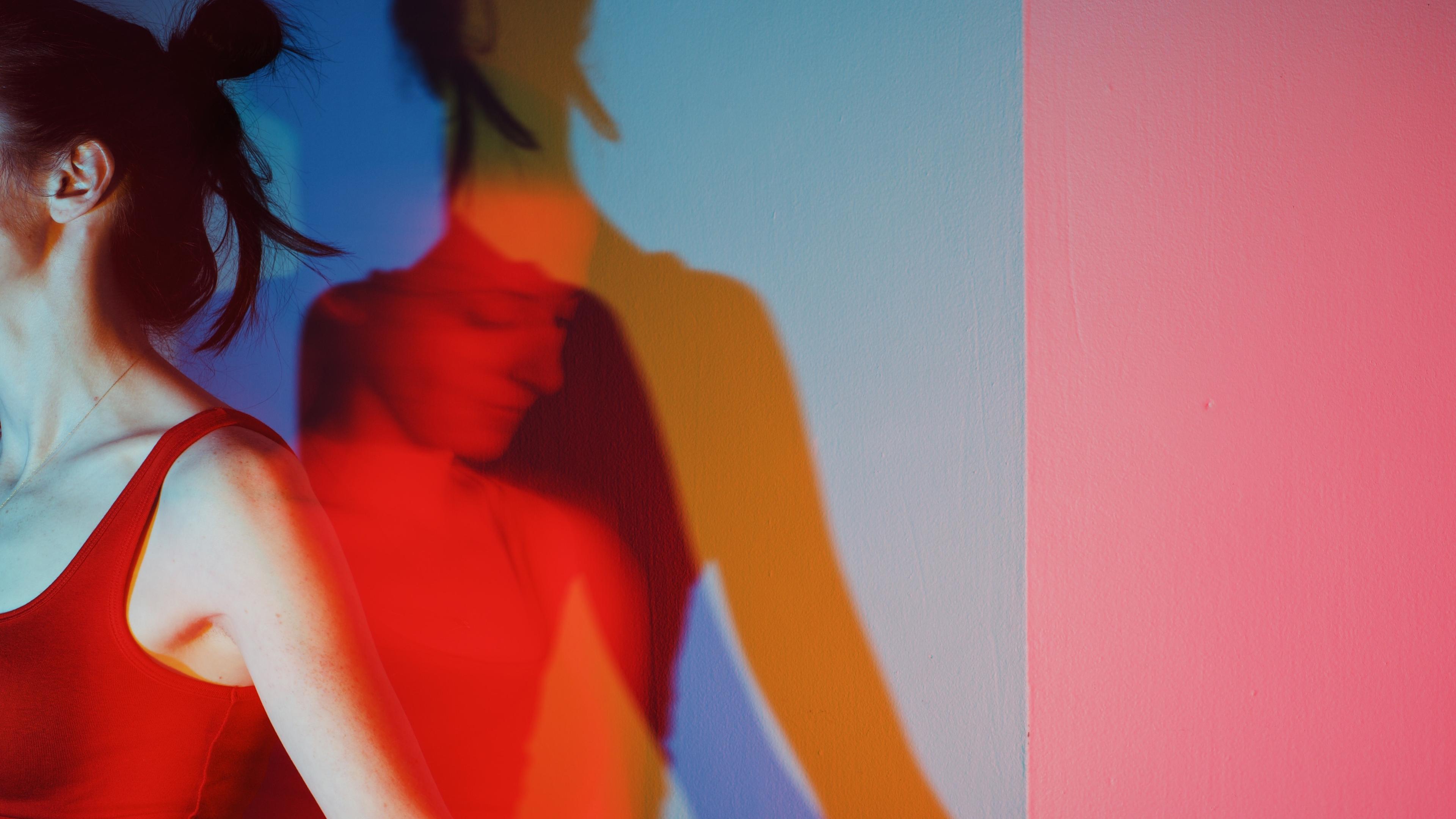 Photo of a woman in a red top with colourful overlapping shadows on a wall displaying red, blue and yellow hues.