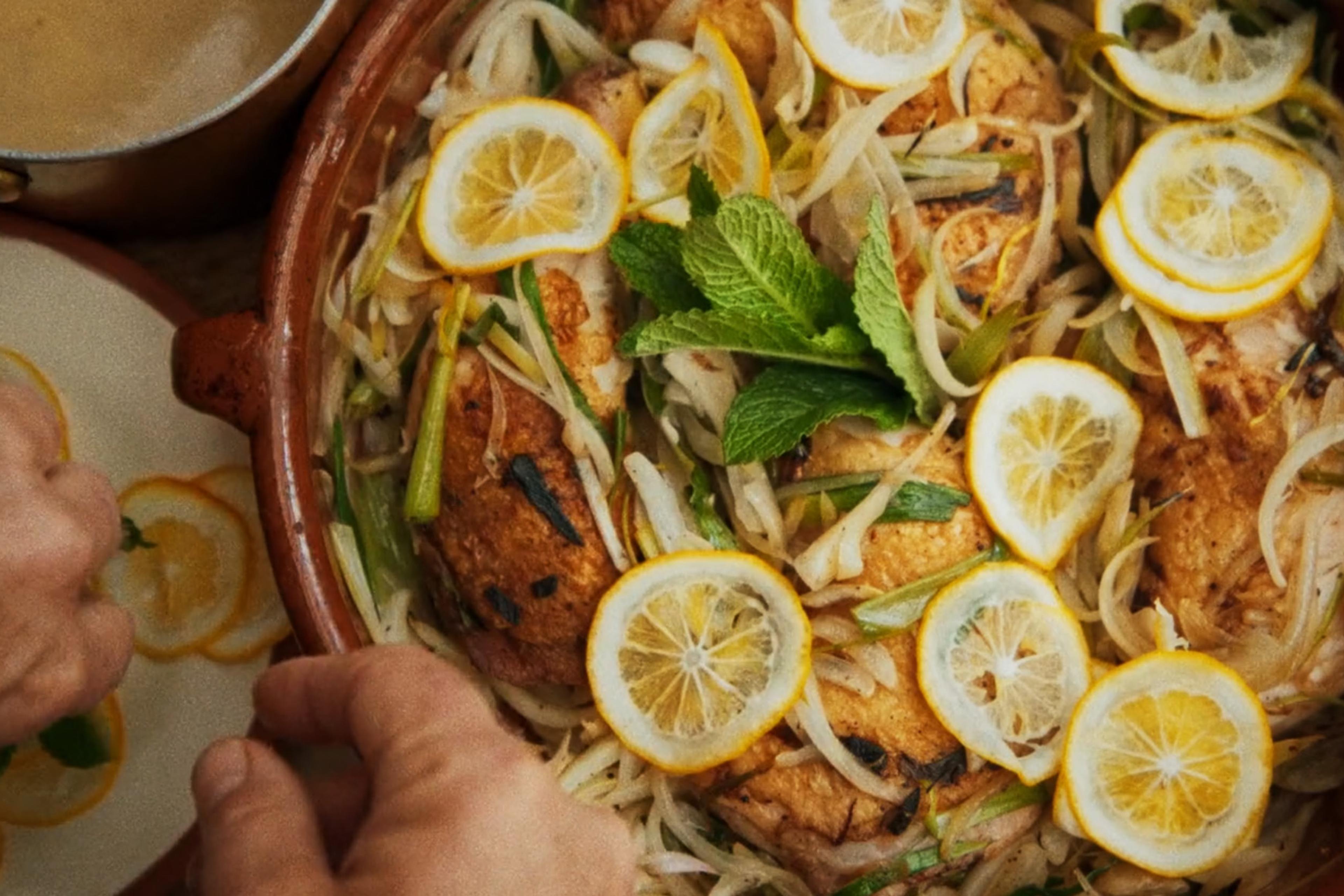 A dish of roasted chicken garnished with lemon slices, bean sprouts, and fresh mint in a clay pot, with a person arranging lemon slices on a nearby plate.