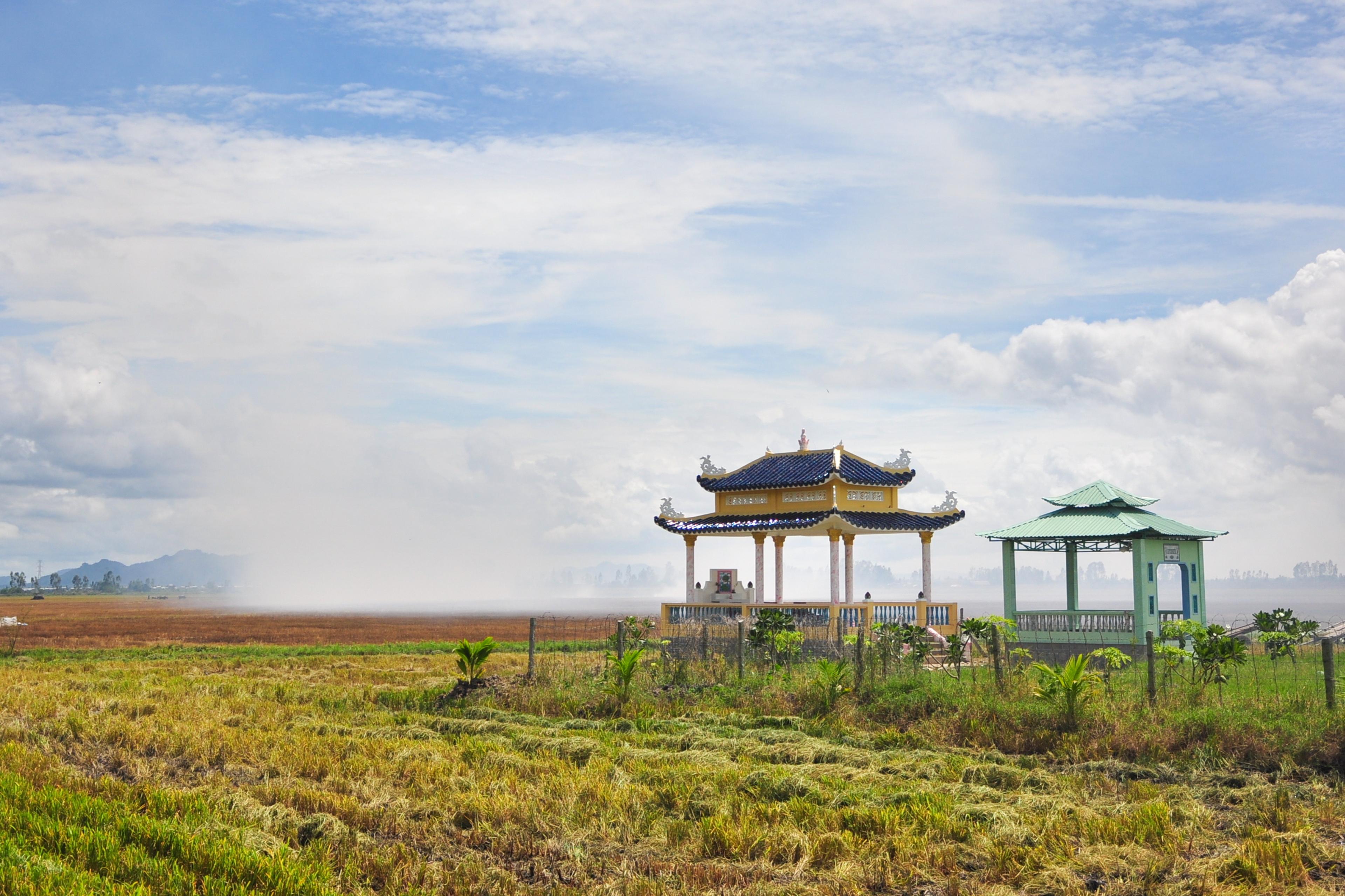 Photo of a rural landscape with two traditional Asian pavilions, green fields and a cloudy sky in the background.
