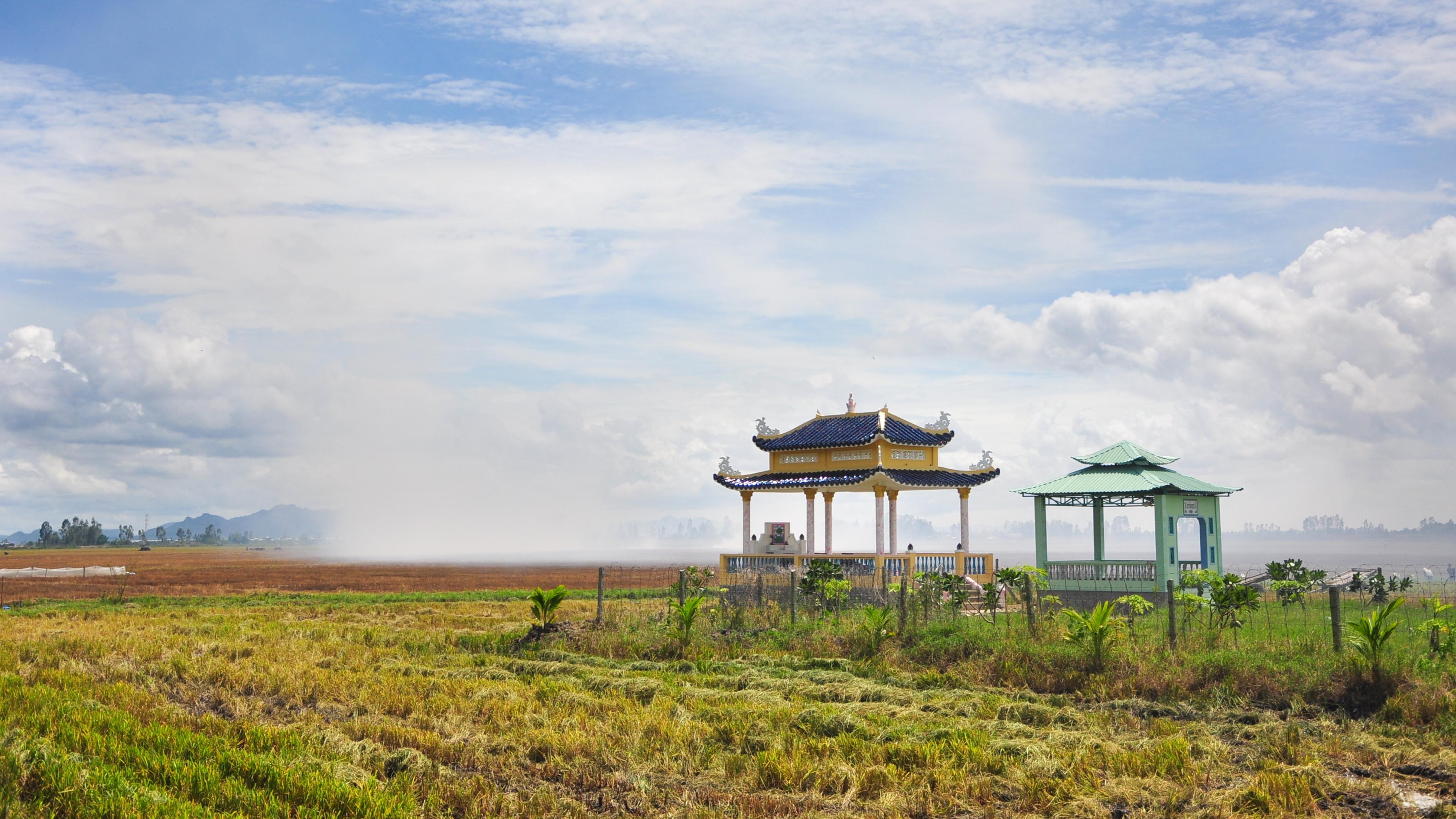 Photo of a rural landscape with two traditional Asian pavilions, green fields and a cloudy sky in the background.