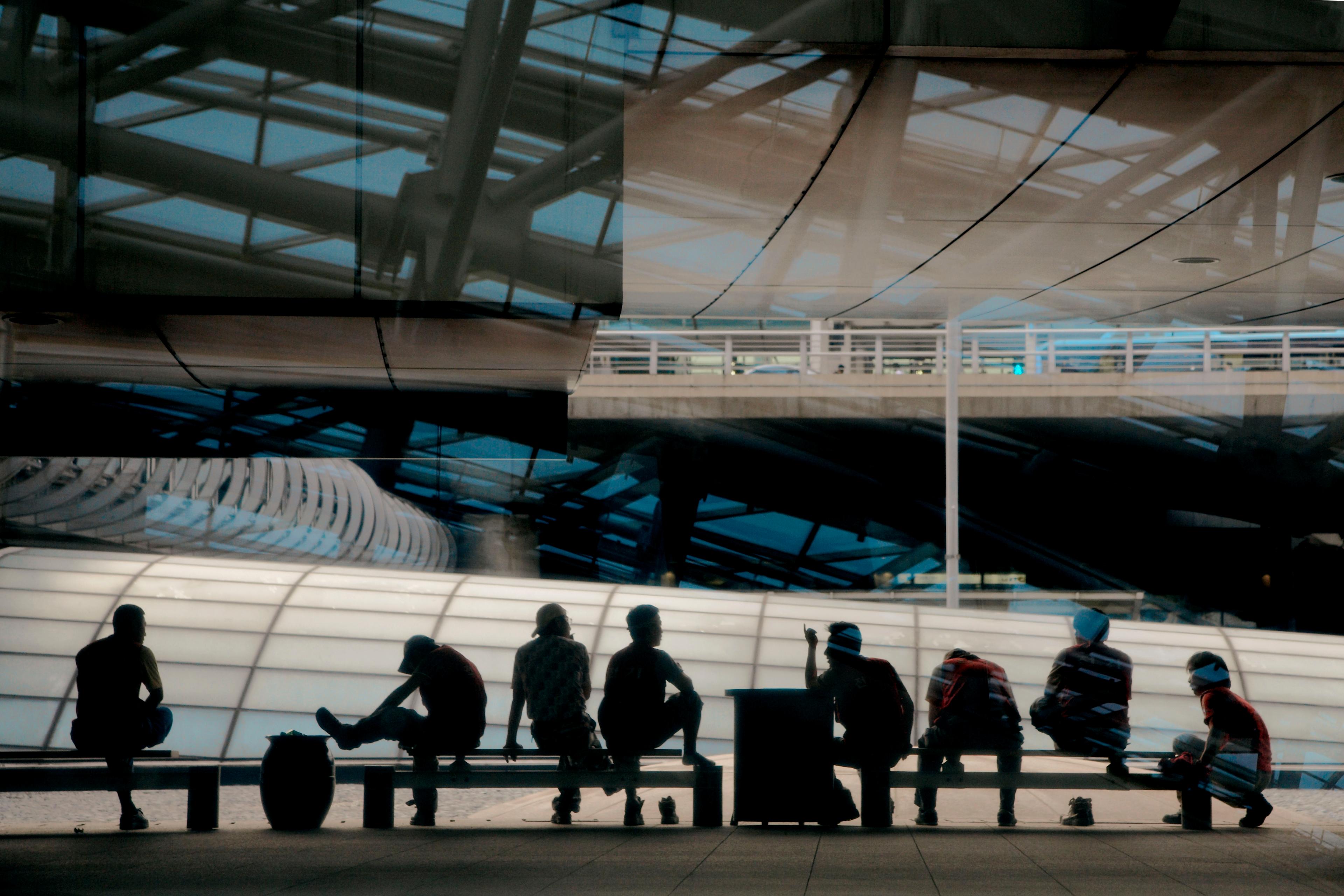 Photo of silhouetted people sitting on benches in a modern building with glass and metal architecture overhead.