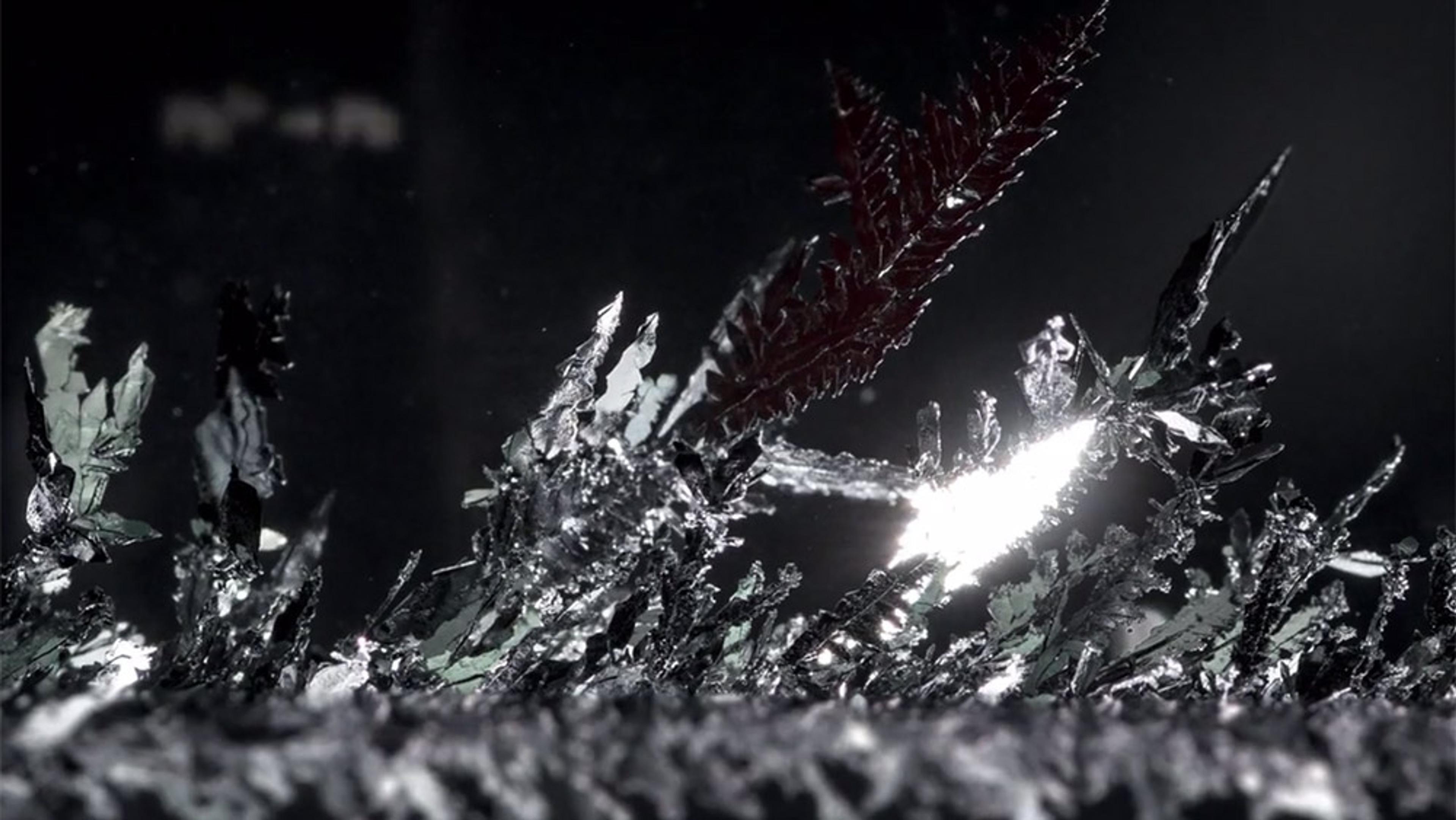 A close-up of intricate shiny metallic crystal formations against a dark background. The crystals reflect light dramatically.