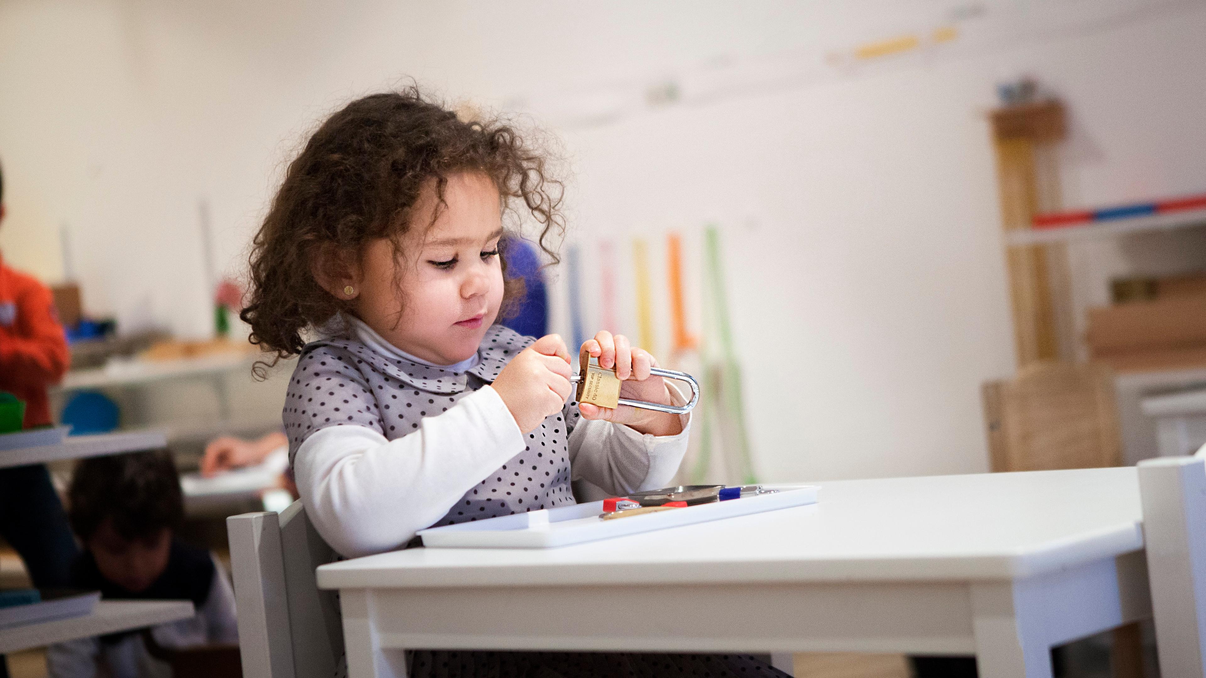 Photo of a young child in a classroom sitting at a table playing with a padlock, with blurred shelves and children in the background.