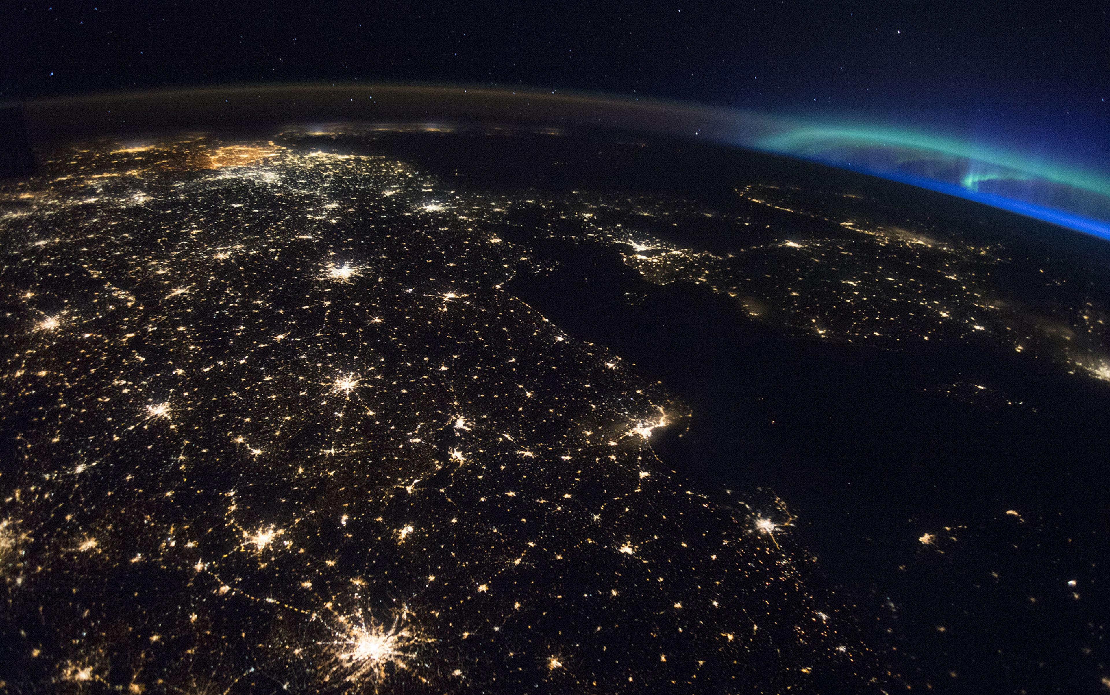 View from space of Europe at night, showing bright city lights and the Northern Lights in the distance. The image features a clear outline of countries and coastlines illuminated by artificial light, with a dark sky speckled with stars overhead.