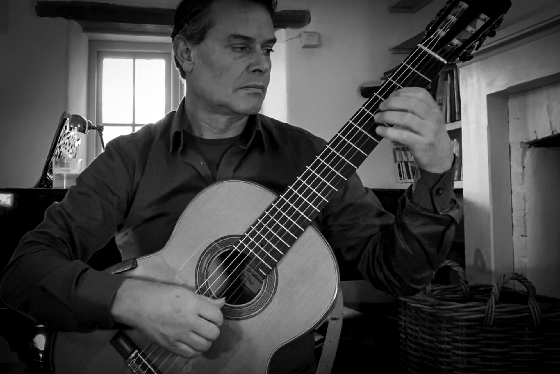 Black and white photo of a man playing a classical guitar in a room with a window in the background.