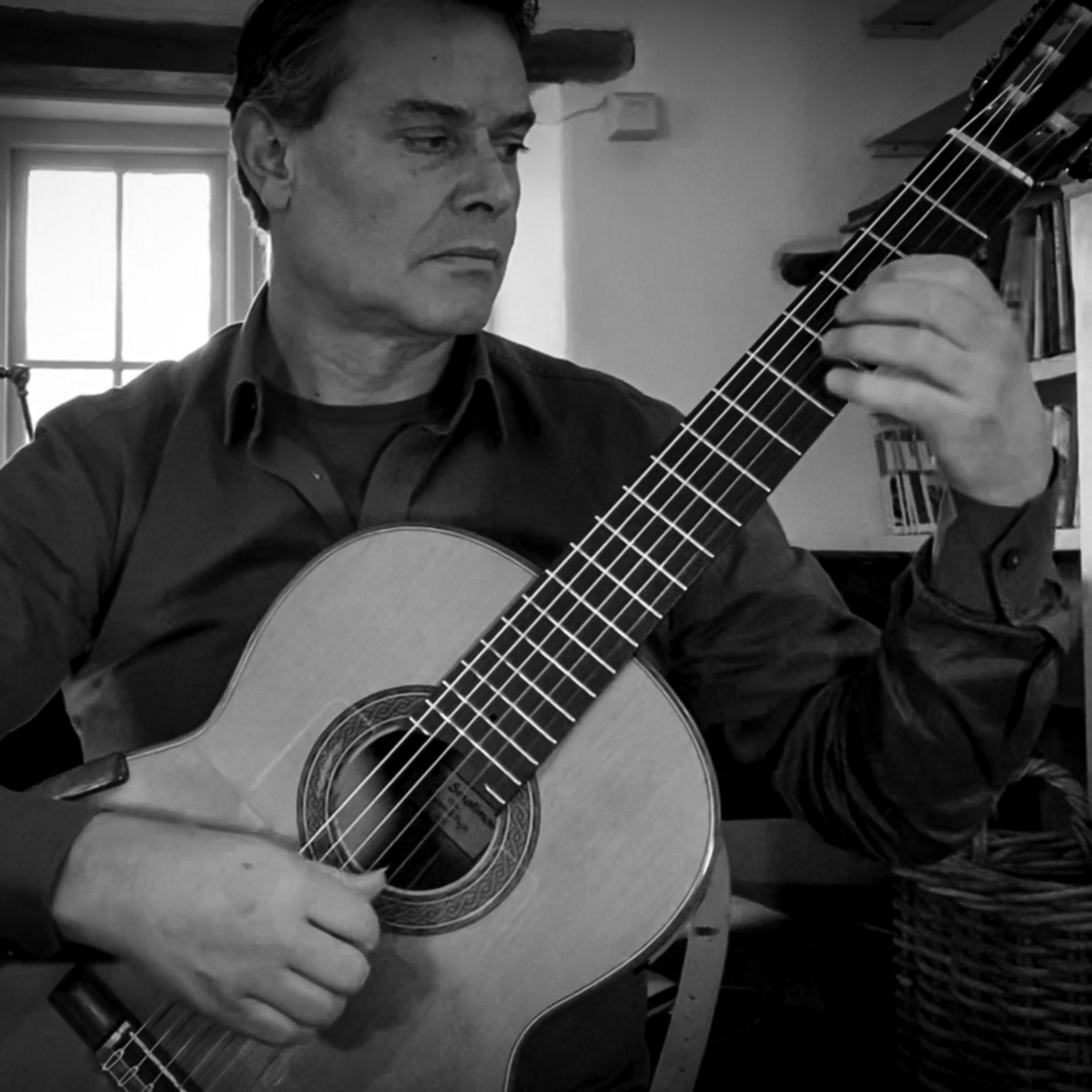 A black-and-white image shows a person playing classical guitar beside a window.