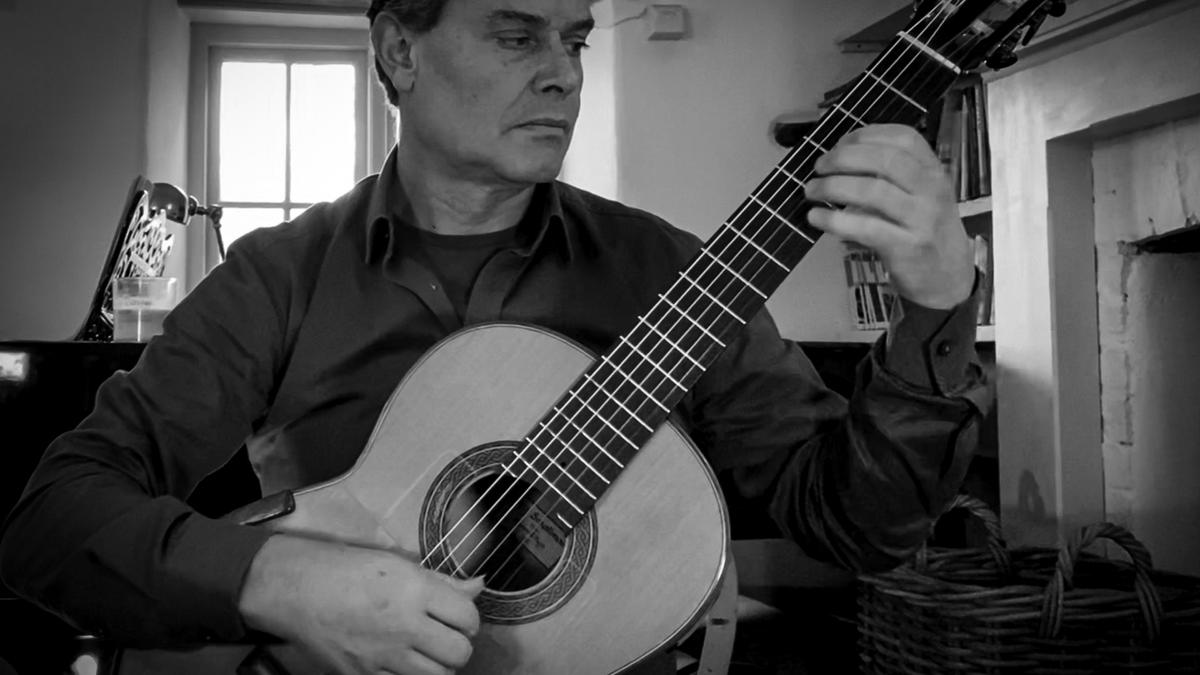 Black and white photo of a man playing a classical guitar in a room with a window in the background.