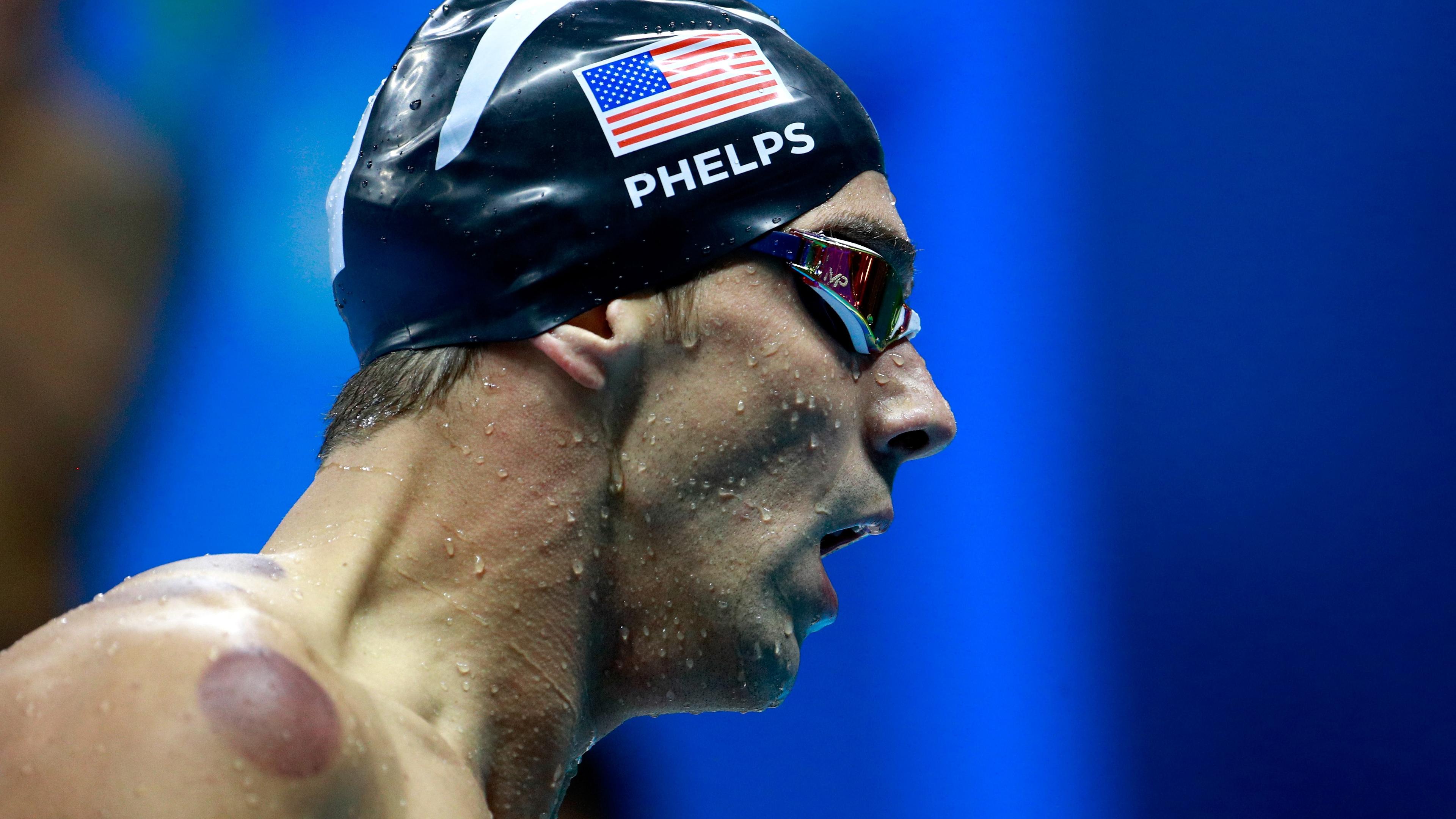 Photo of a swimmer with goggles and cap, water droplets visible on skin, seen in profile against a blue background.