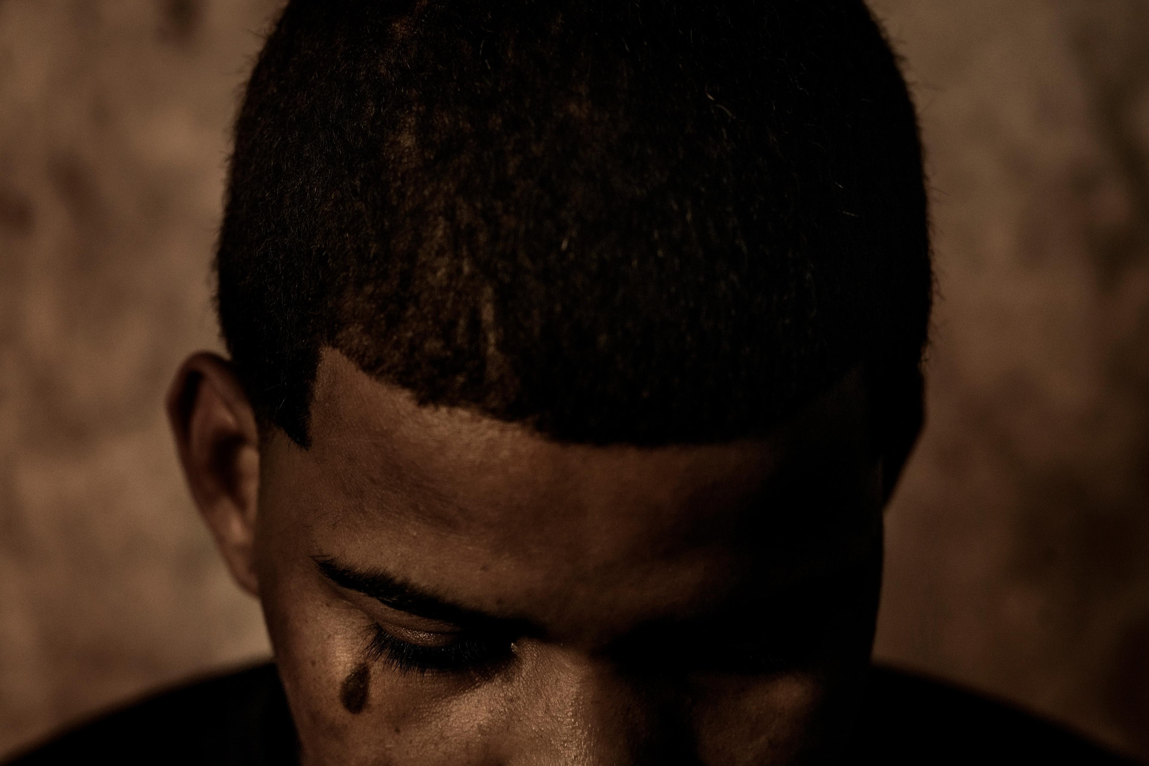A monochrome photo of a person with a shaved head looking down in low light, a tattooed teardrop visible near their eye.