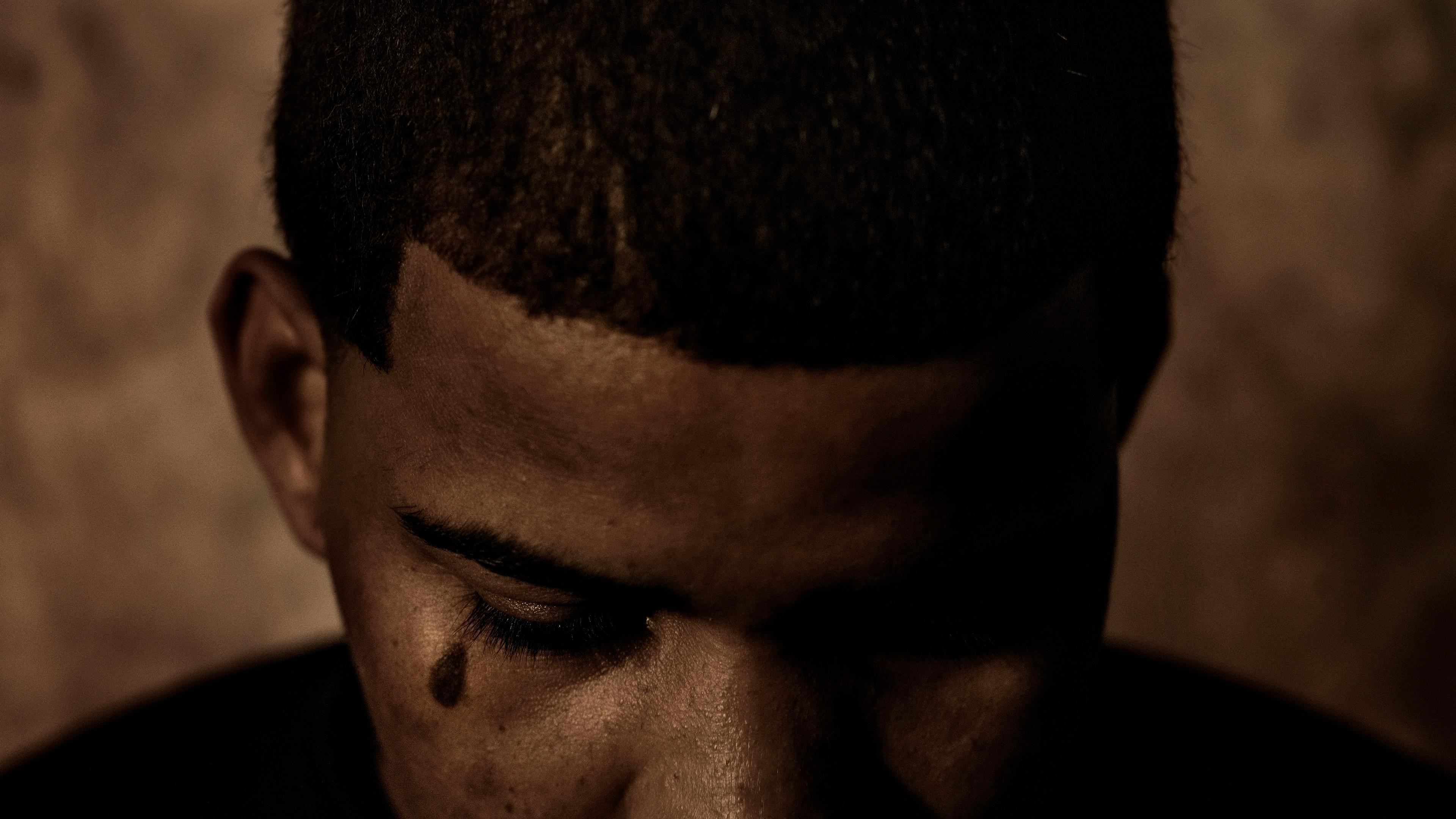 A monochrome photo of a person with a shaved head looking down in low light, a tattooed teardrop visible near their eye.