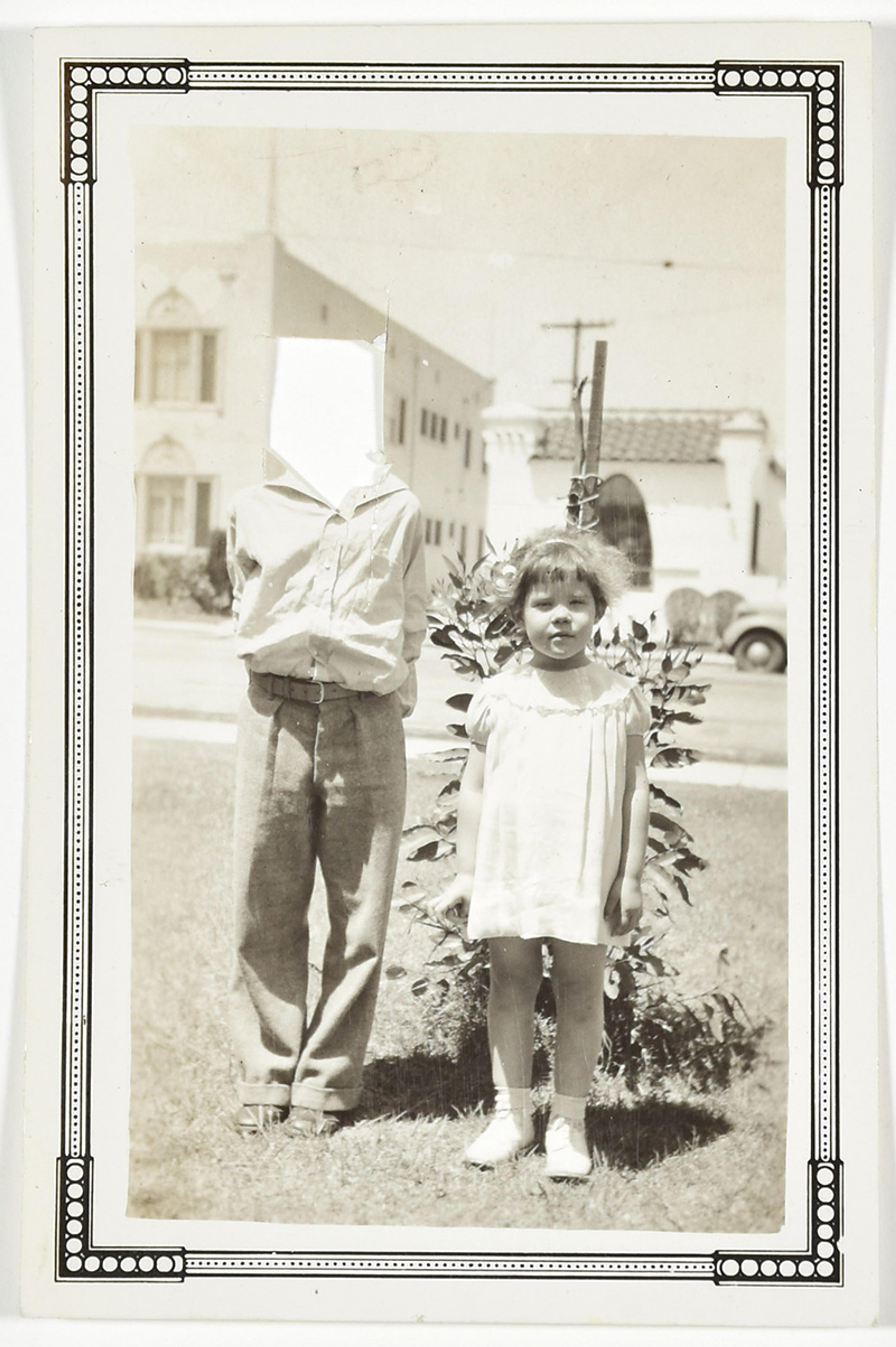 A black-and-white photo of a young girl standing next to a boy whose face is obscured in front of a plant, outside.