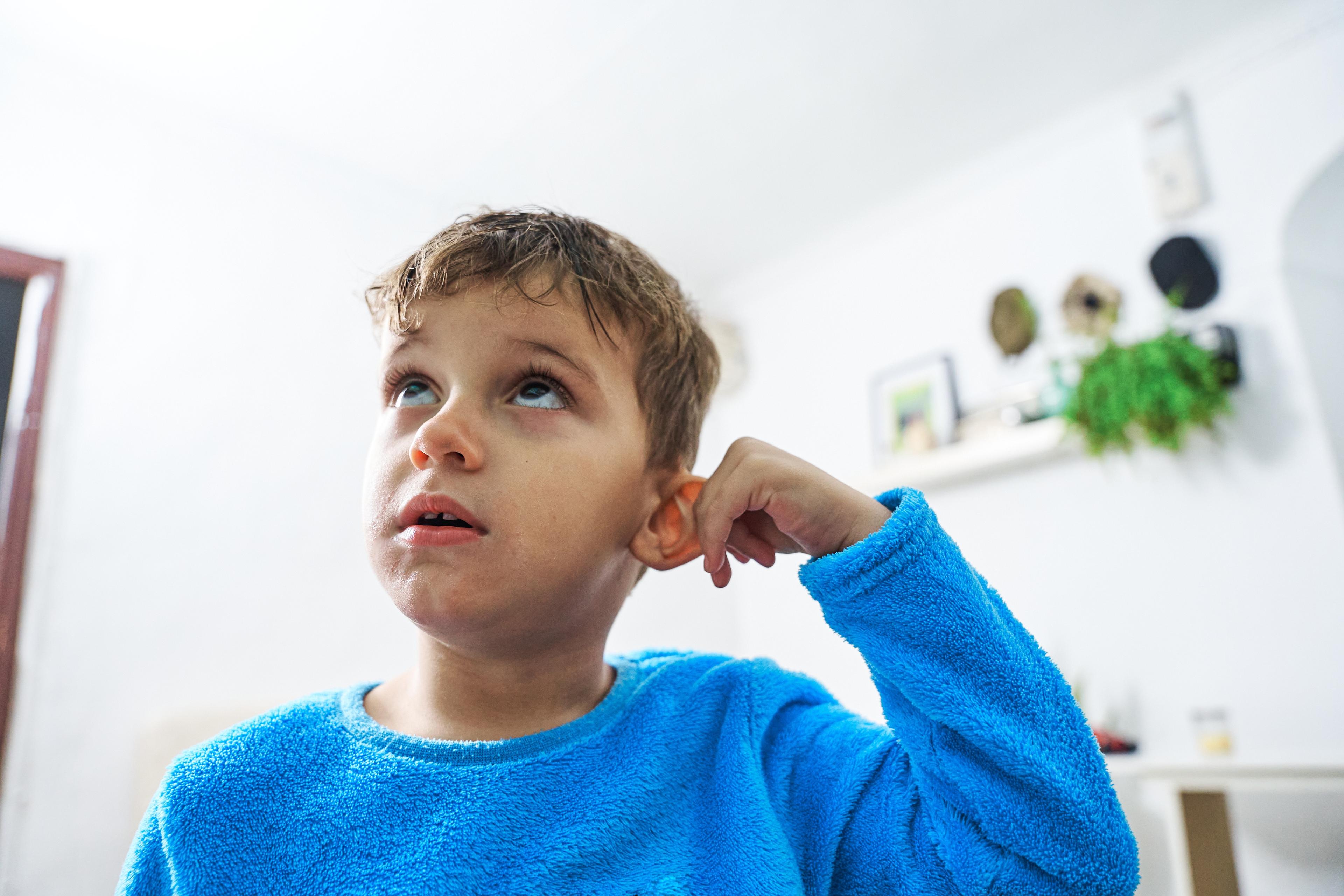 Photo of a young boy in a blue jumper looking up thoughtfully with a finger touching his ear in a bright room.