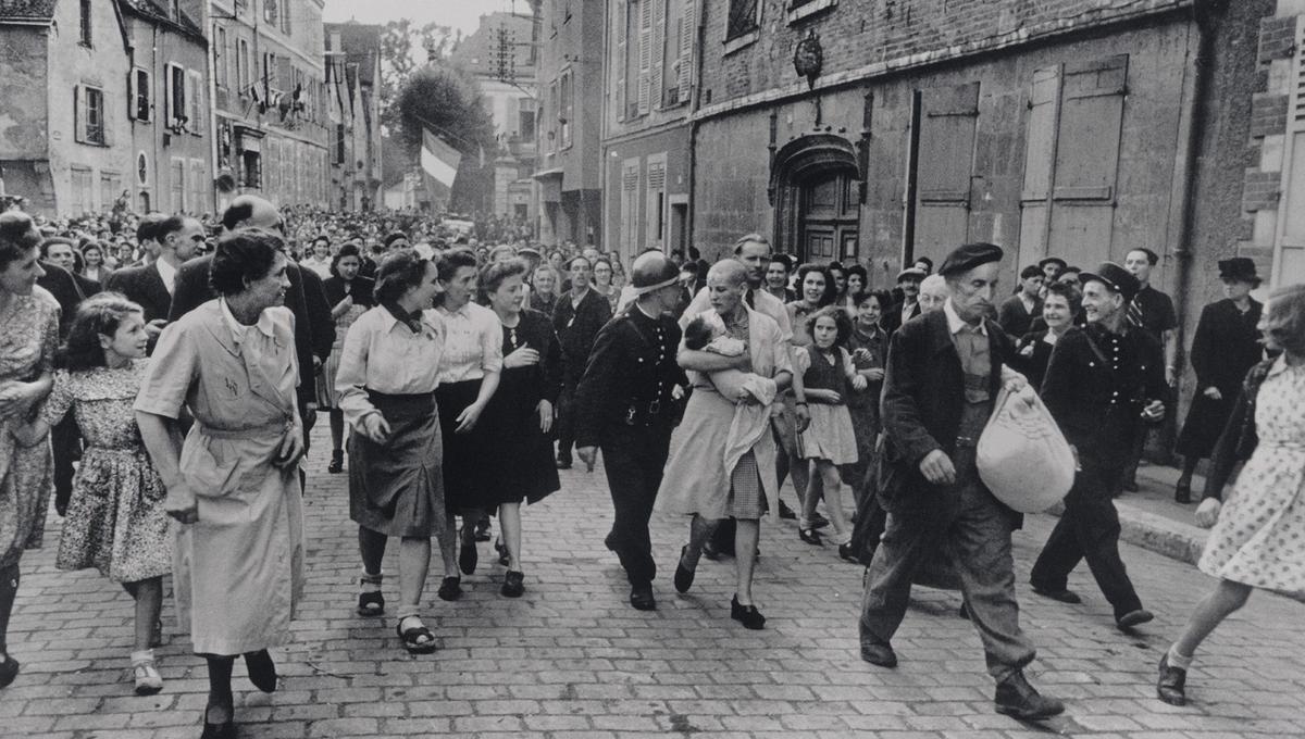 An angry mob of people jeer at a woman carrying a young baby up a cobbled street. Her head is shaved