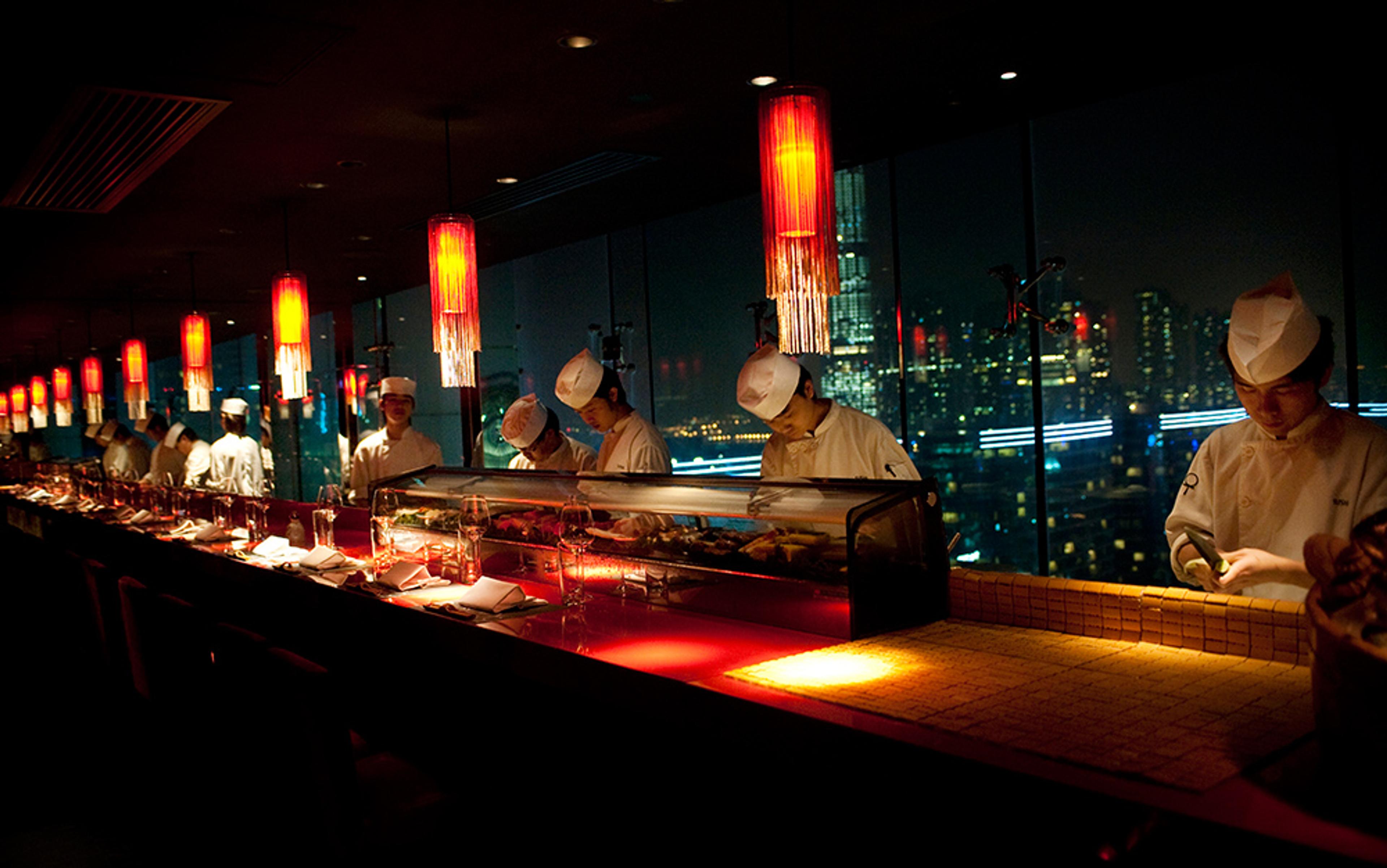 Chefs preparing food behind a sushi bar in a dimly lit restaurant with red lamps and a city skyline visible through windows.