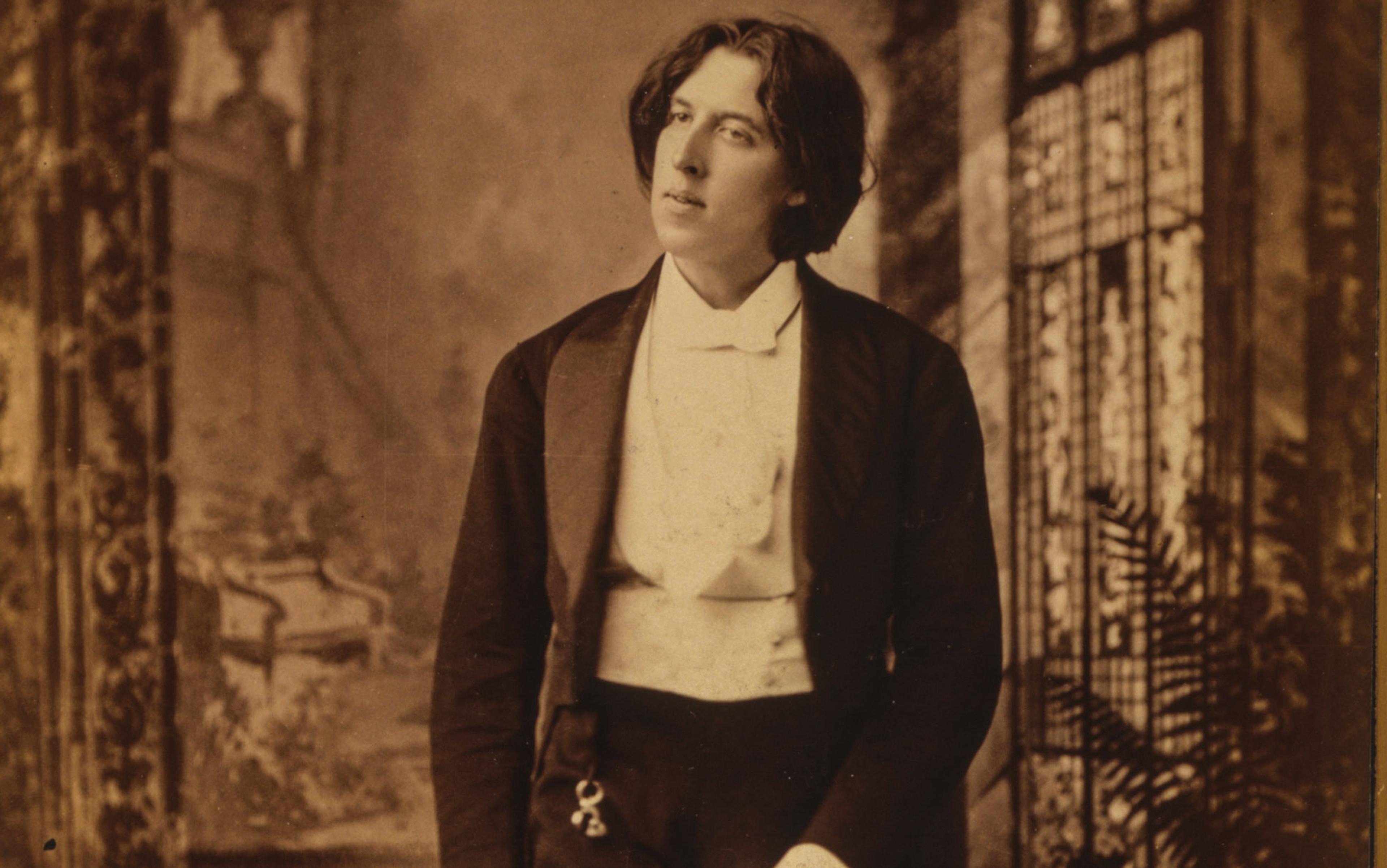 Sepia-toned photo of a person in formal Victorian attire, standing with a slightly tilted head, against an ornate backdrop.