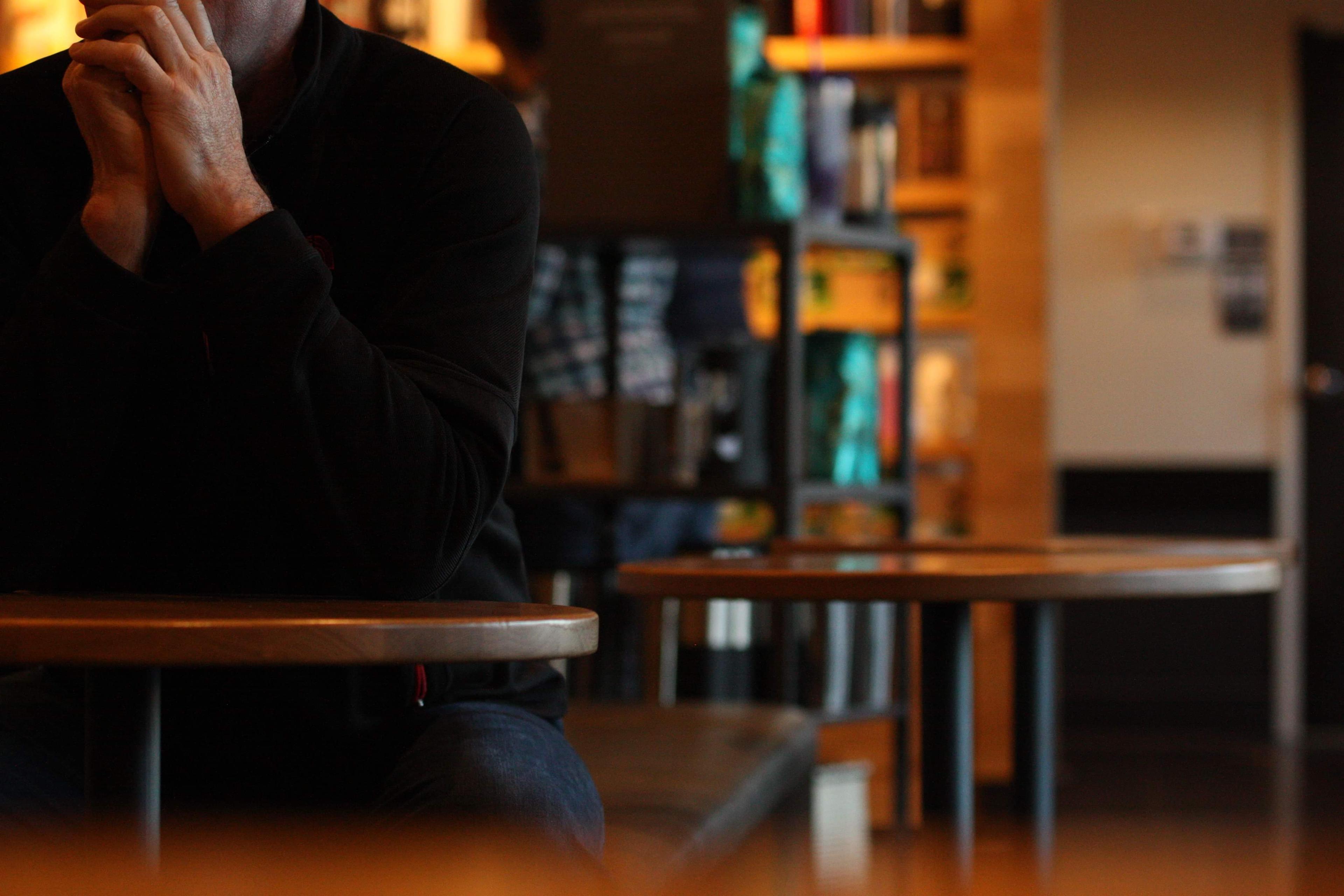 A photo of a person sitting at a table in a cosy café setting. The background is softly blurred, showing shelves and decor.
