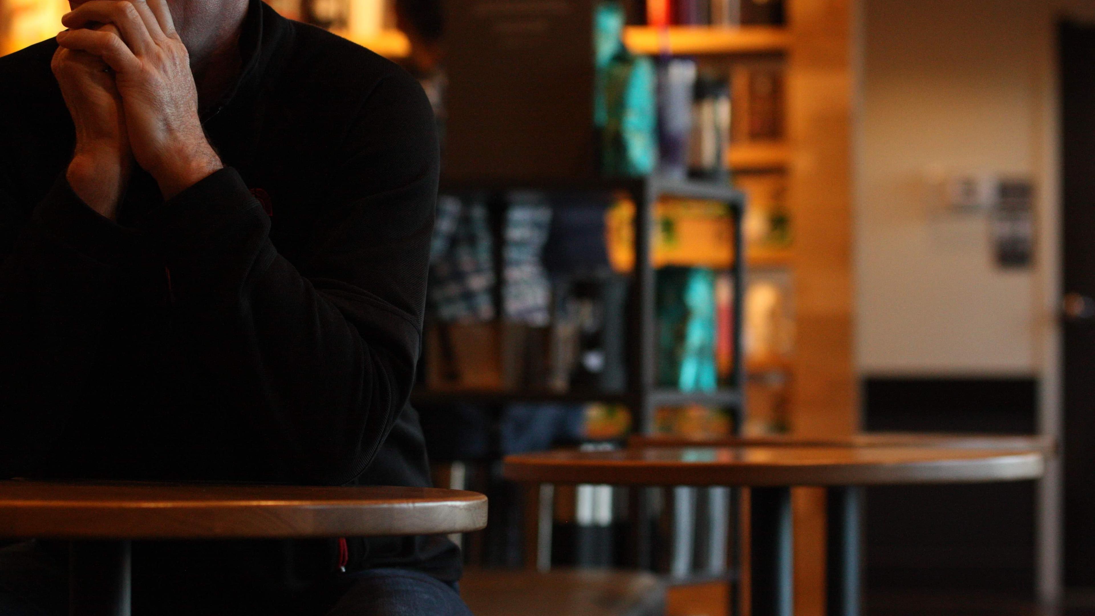A photo of a person sitting at a table in a cosy café setting. The background is softly blurred, showing shelves and decor.