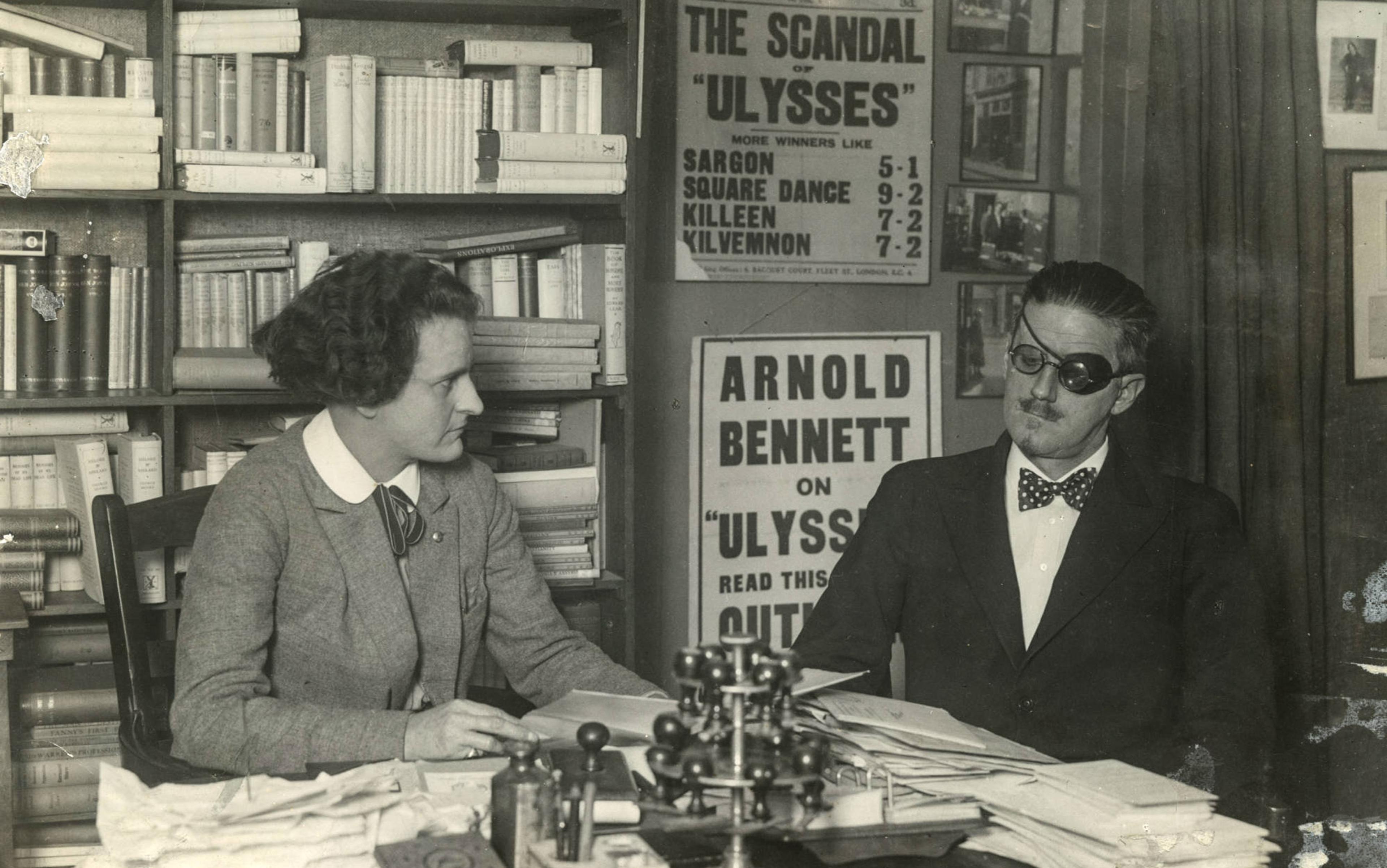 Two people sit at a desk covered in papers. Background includes book shelves and posters about the “Ulysses” novel and Arnold Bennett.