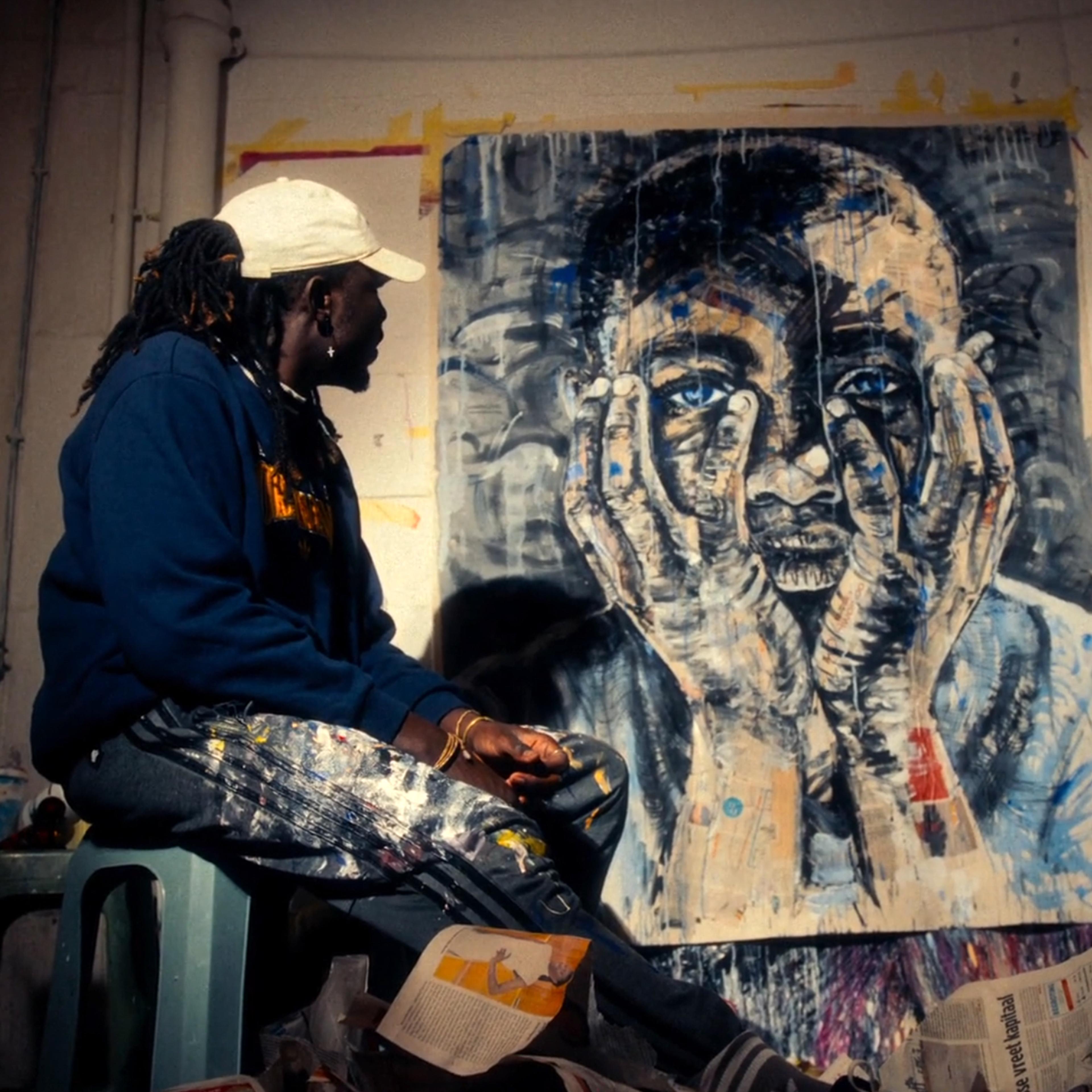 A photo of an artist in a cap and paint-splattered trousers sitting next to a large painting of a man holding his face in his hands.