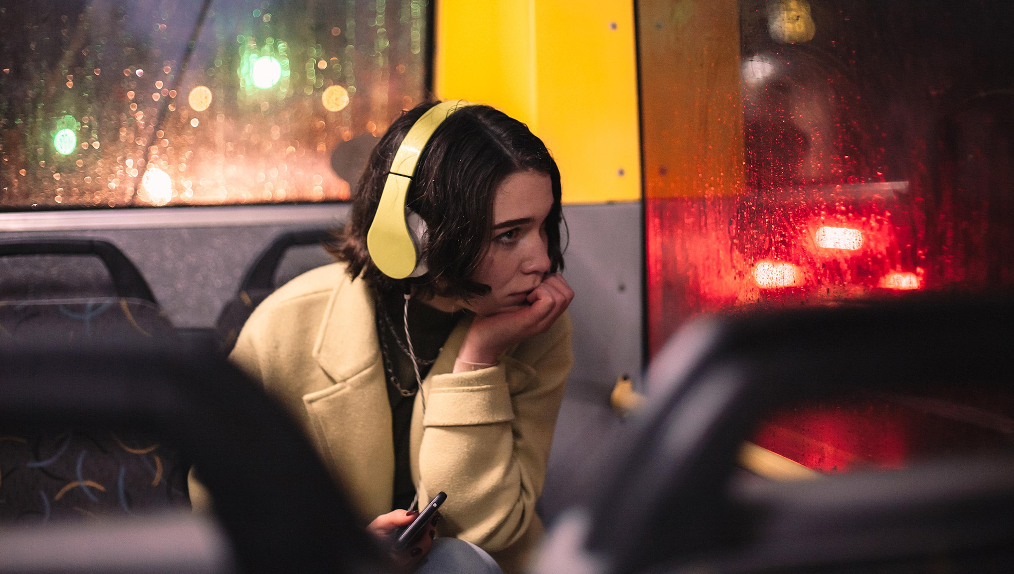 A pensive-looking woman is listening to headphones on a bus on a rainy evening