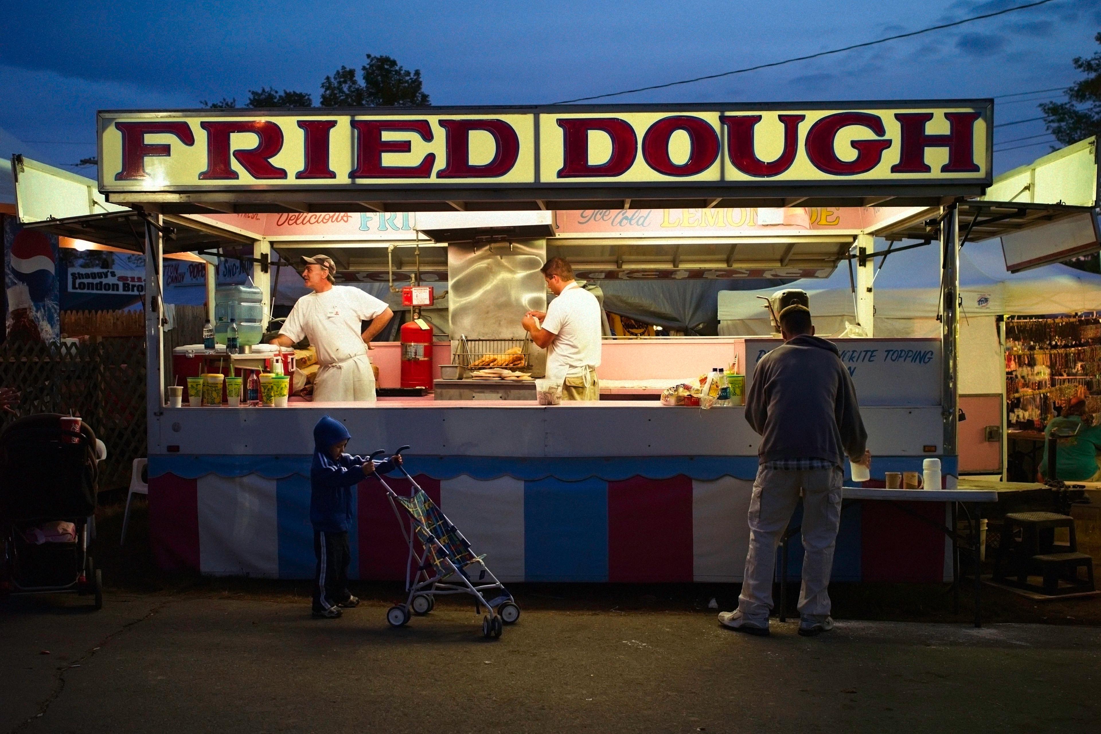 Photo of a fried dough stall at a fair with vendors inside, a child with a stroller, and a customer at the counter.
