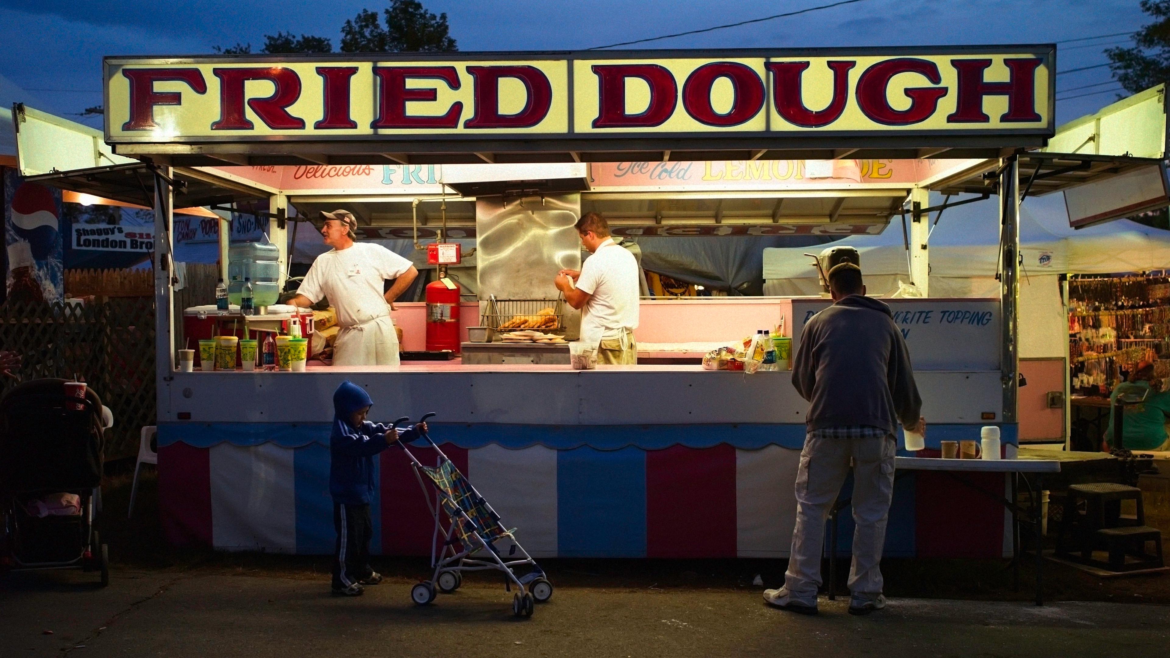 Photo of a fried dough stall at a fair with vendors inside, a child with a stroller, and a customer at the counter.