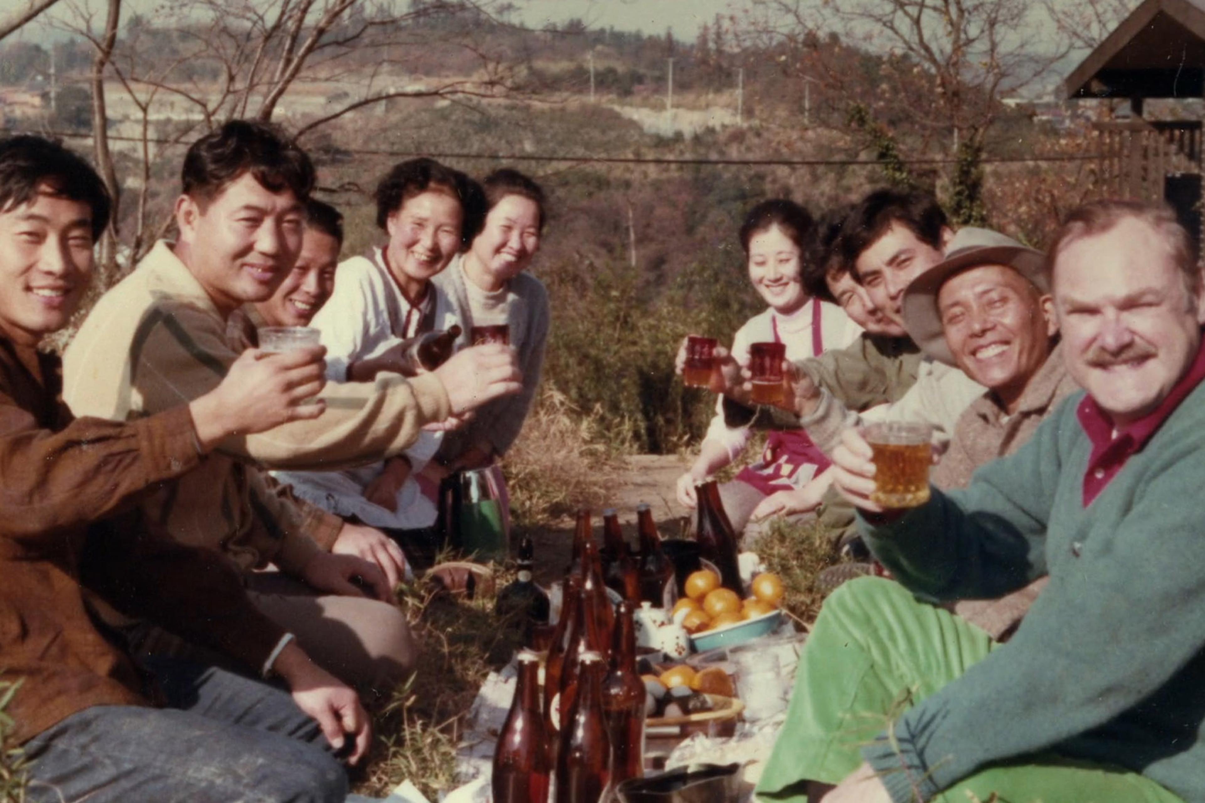 Photo of a group of people sitting outside, smiling and raising drinks together. A picnic with bottles and fruits is visible in front.