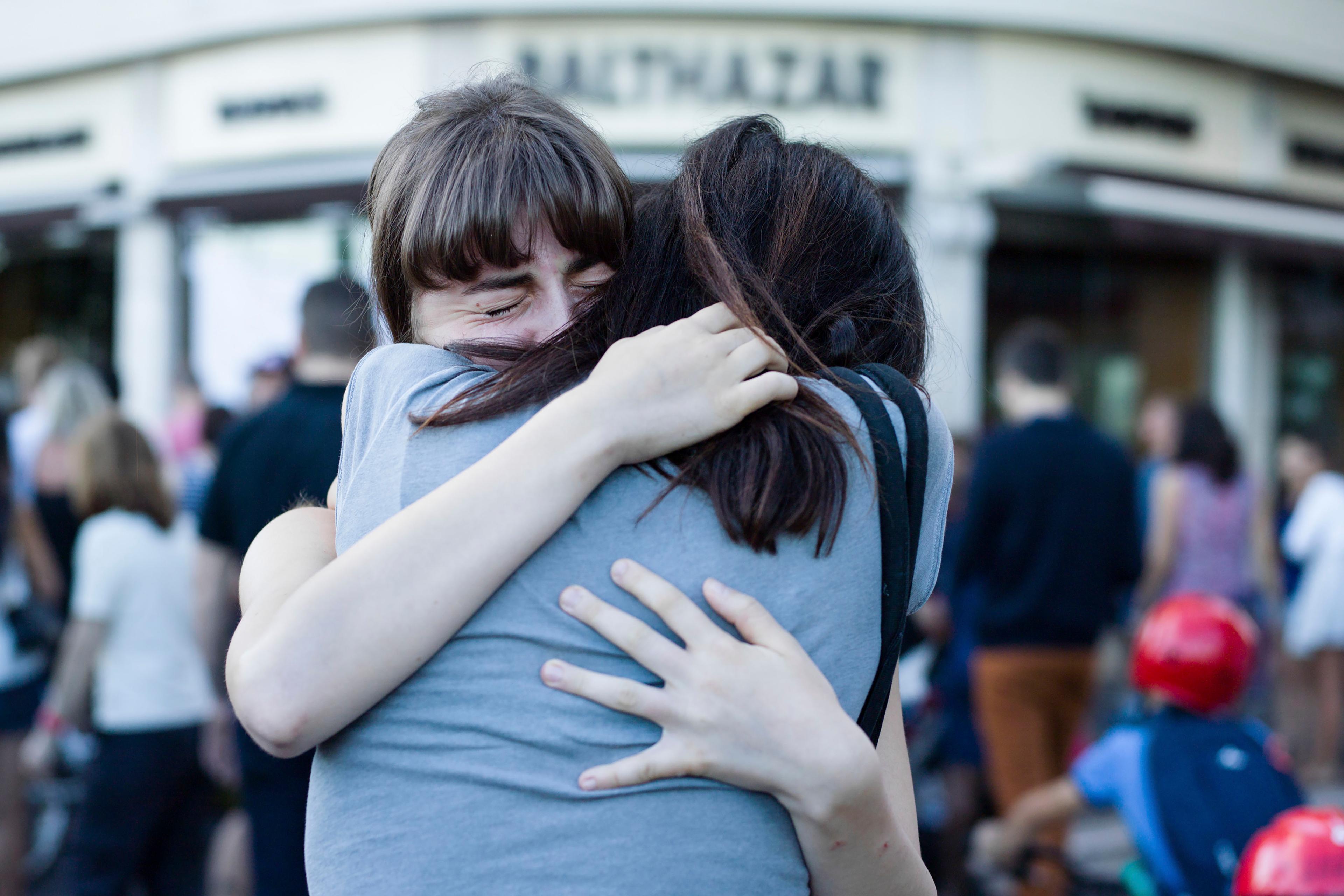 Photo of two people embracing tightly outdoors with eyes closed, surrounded by a blurred crowd in the background.