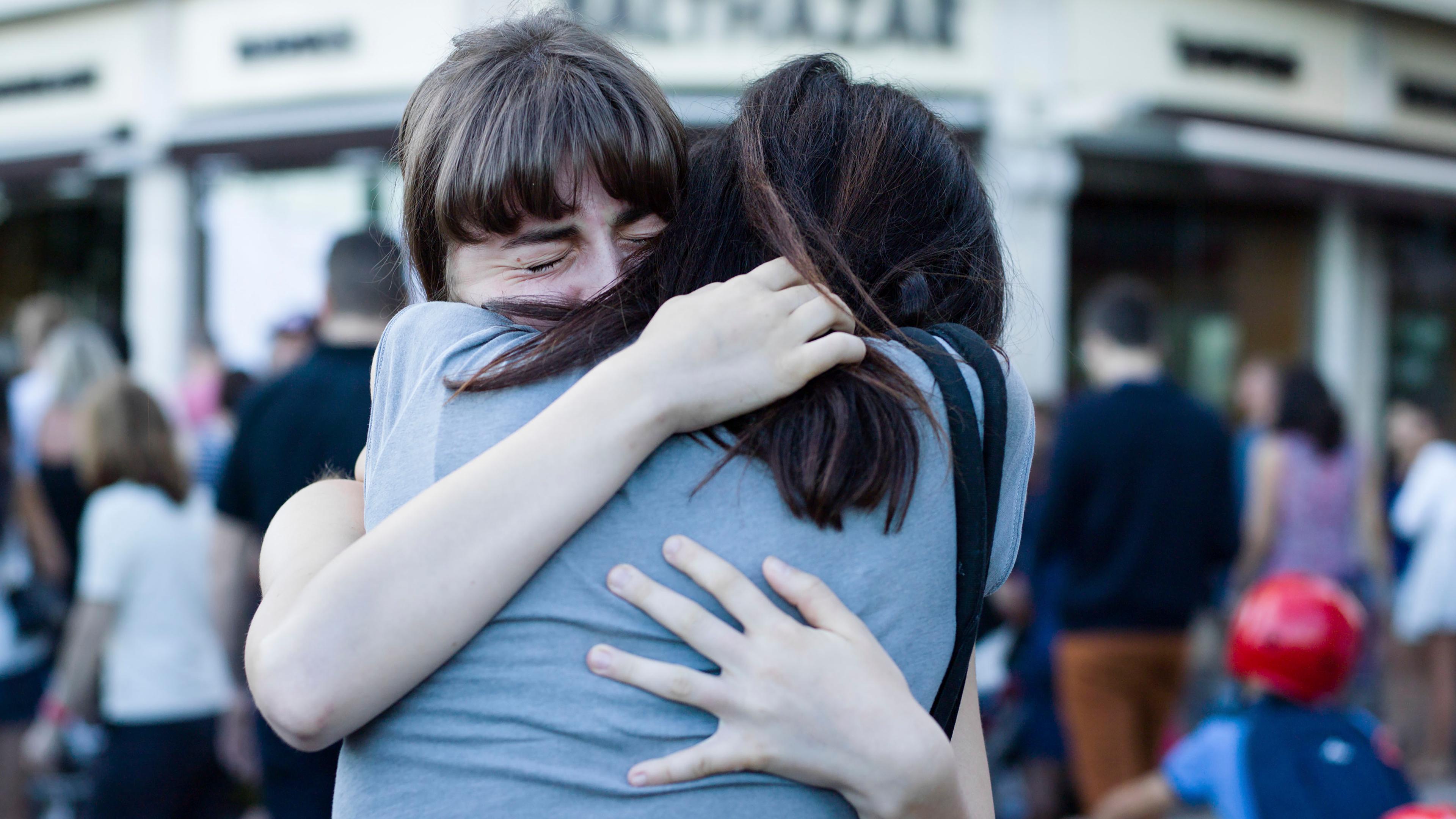 Photo of two people embracing tightly outdoors with eyes closed, surrounded by a blurred crowd in the background.