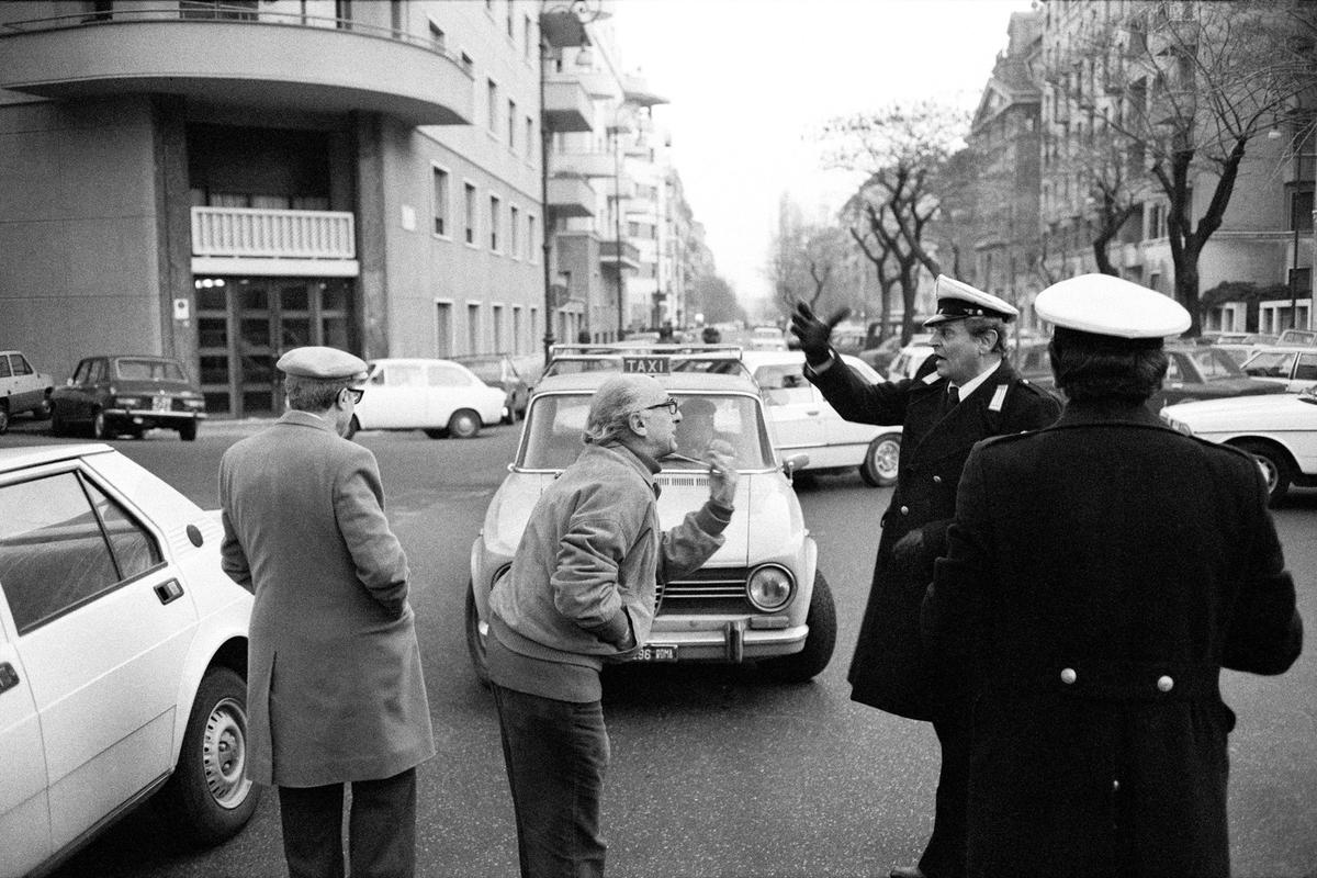 Black and white photo of a street scene with a heated discussion involving a traffic officer and men near cars.