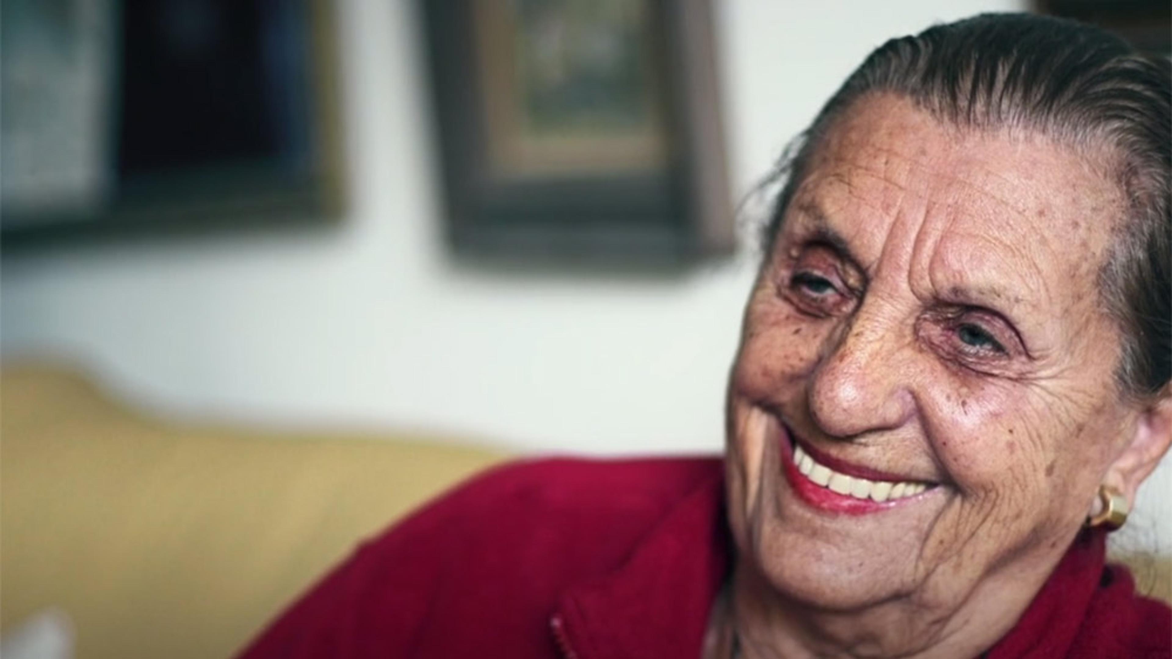 An elderly woman with a warm smile wearing a red top, sitting indoors with framed pictures on the wall in the background.
