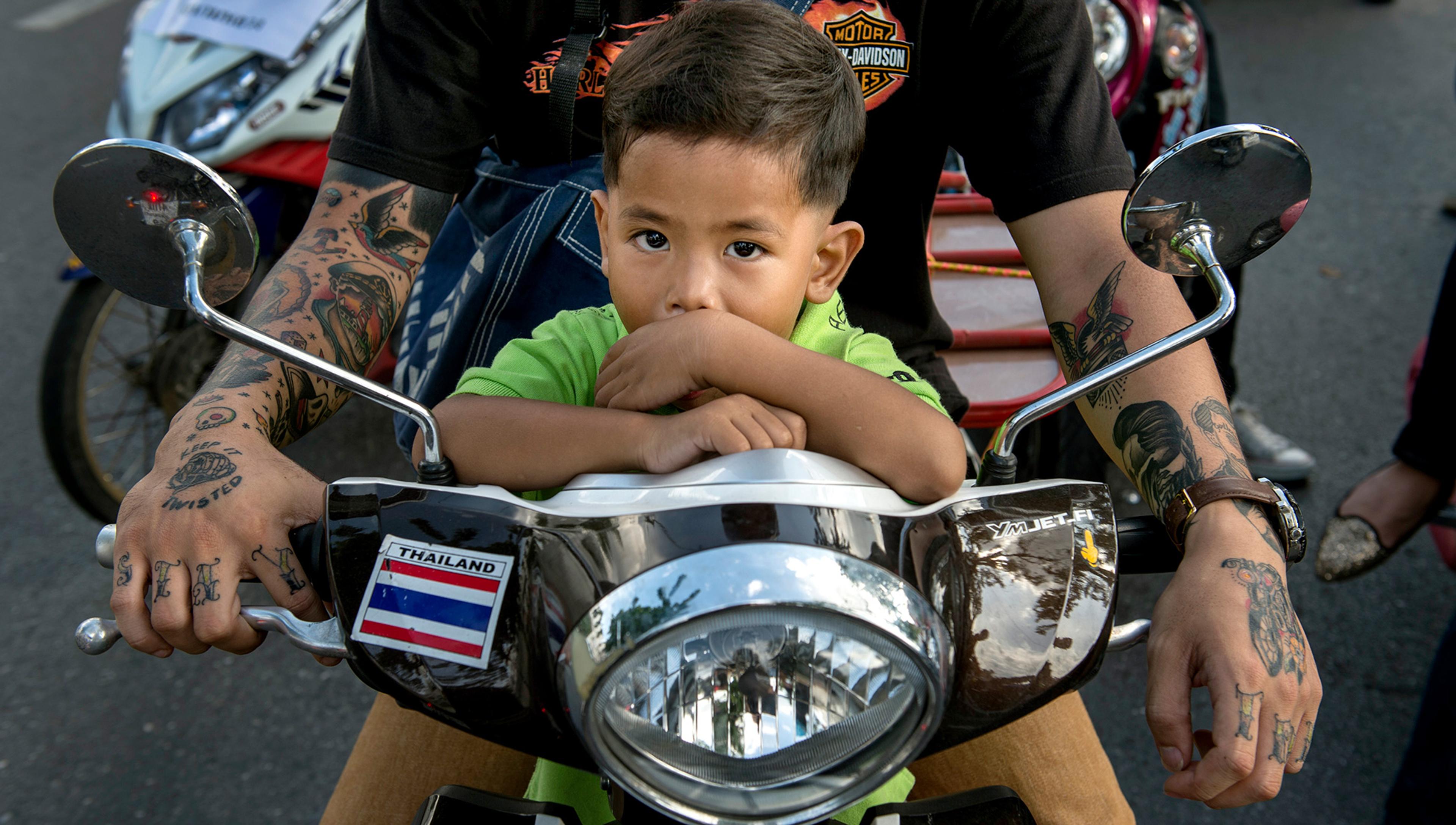 A young child passenger sits on the front of a motor scooter, with an older male rider partly visible behind. The rider has tattooed arms and a sign on the scooter reads ‘Thailand’