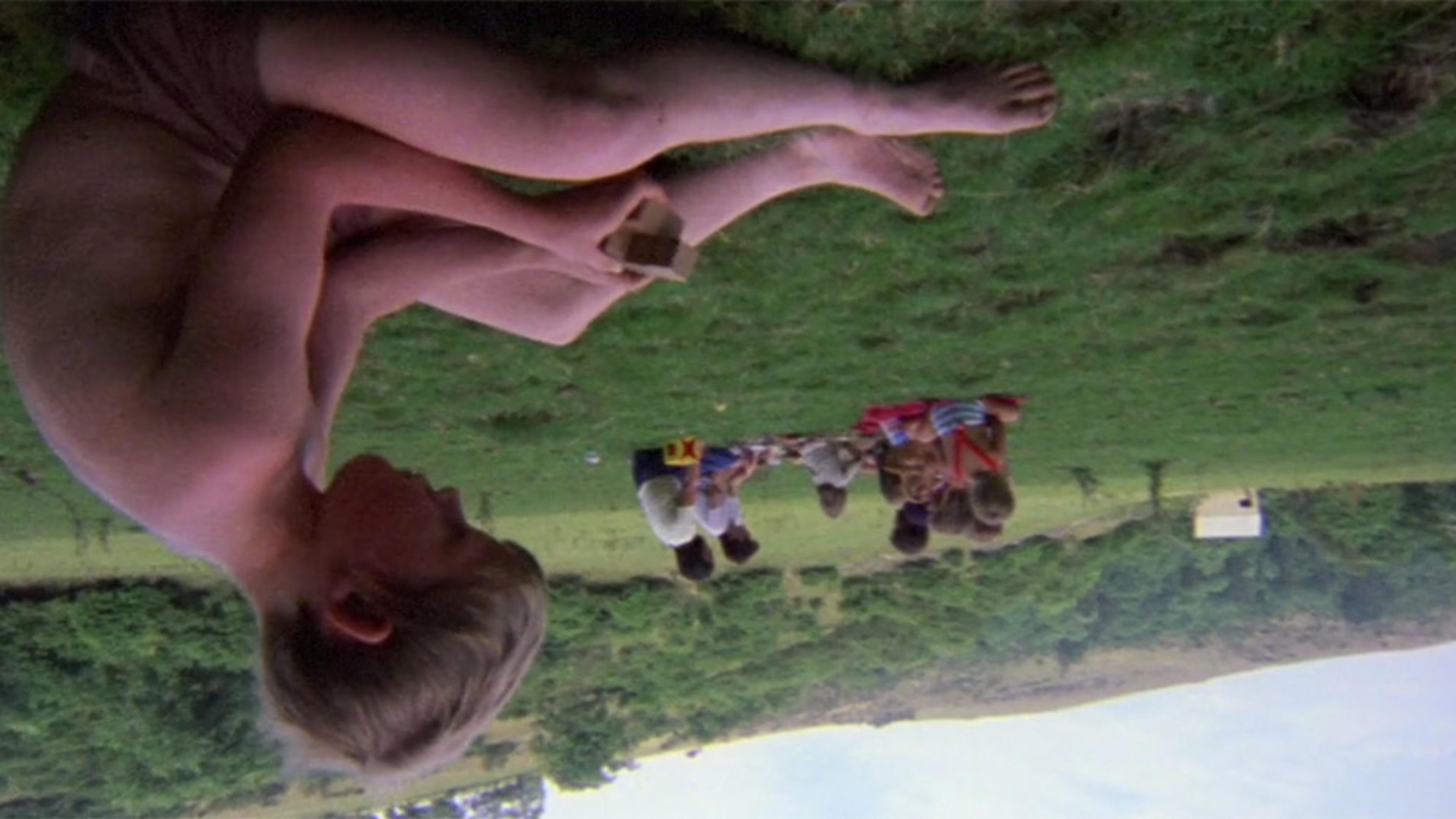 Upside-down photo of a boy sitting on grass holding an object with a group of children in the background in a green, grassy landscape.