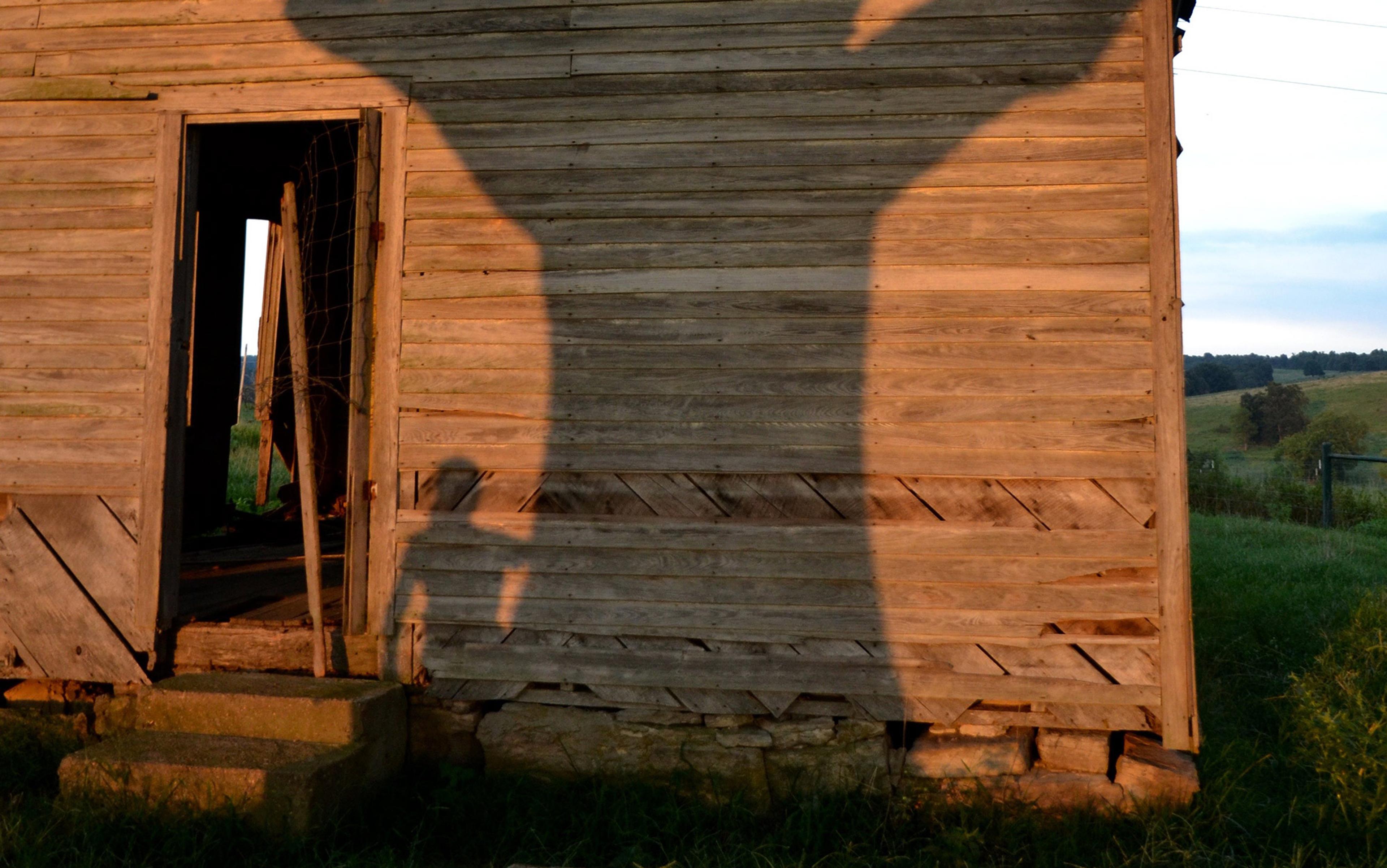 A weathered wooden building with an open door at sunset. Shadows of a tree and a person are cast on the wall.
