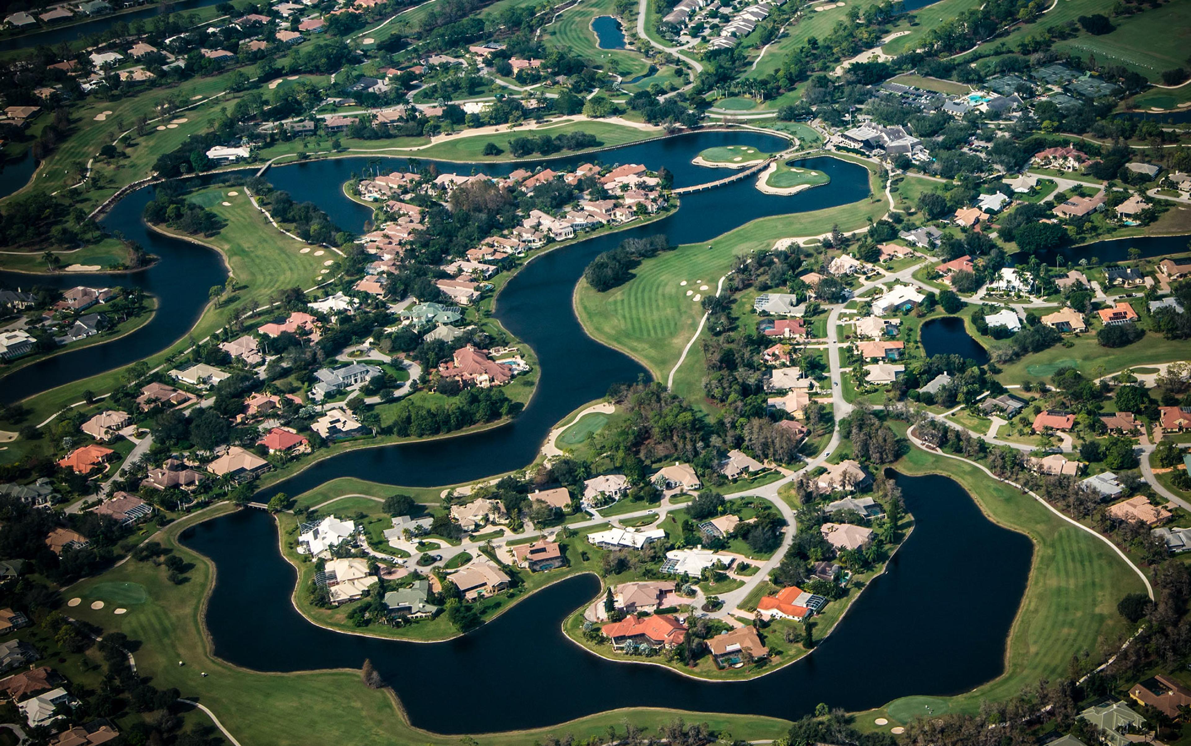 Aerial photo of a residential area with numerous houses, winding roads, small lakes and surrounded by green fields.