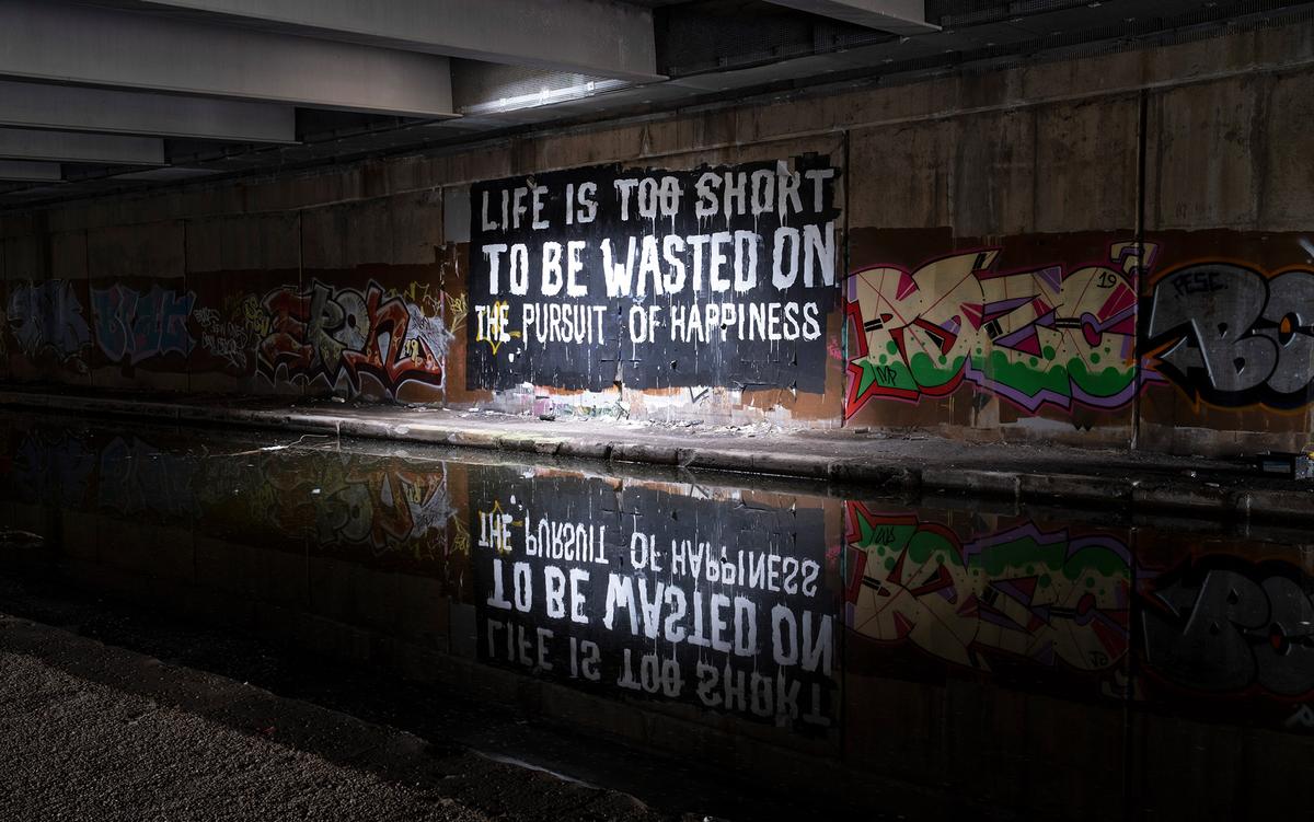 A graffitti sign in an underpass reads ‘Life is too short to be wasted on the pursuit of happiness’; it is reflected in pooling water underneath