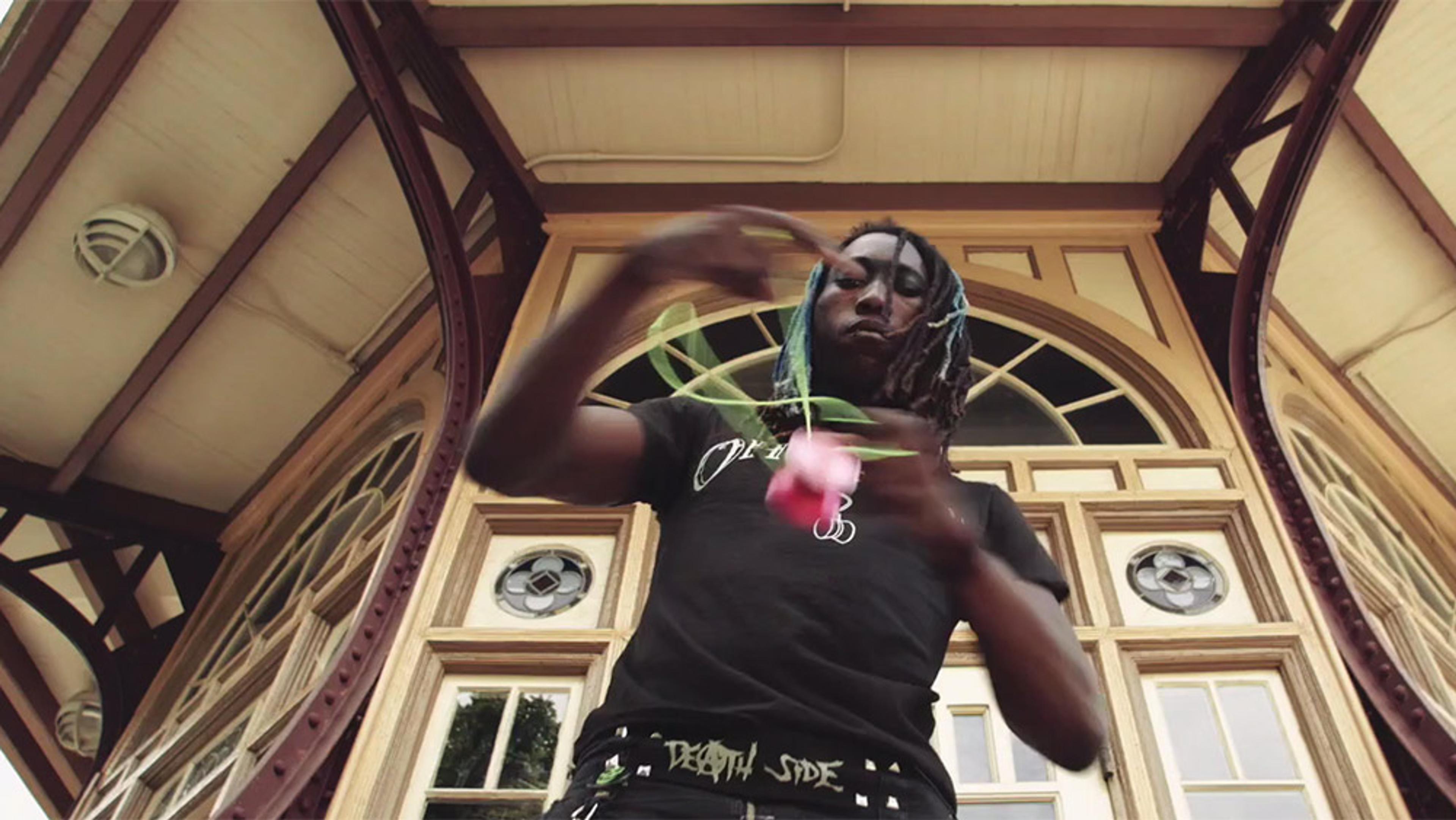A Black man with braided hair playing with a yo-yo in front of a historical-looking building with arched windows and wooden frames.