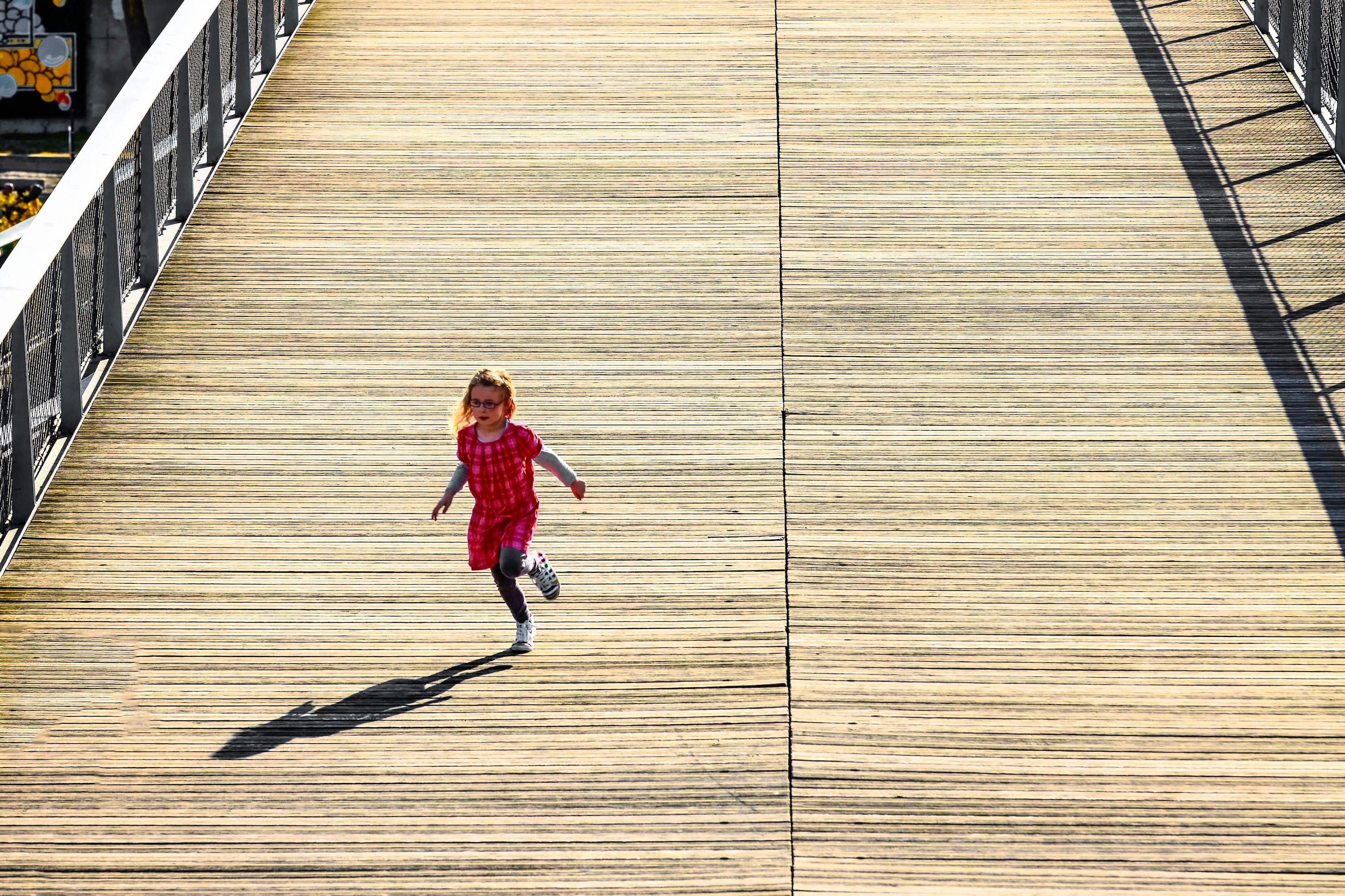 A photo of a child in a red dress running on a wooden bridge with adults walking in the background under a clear sky.