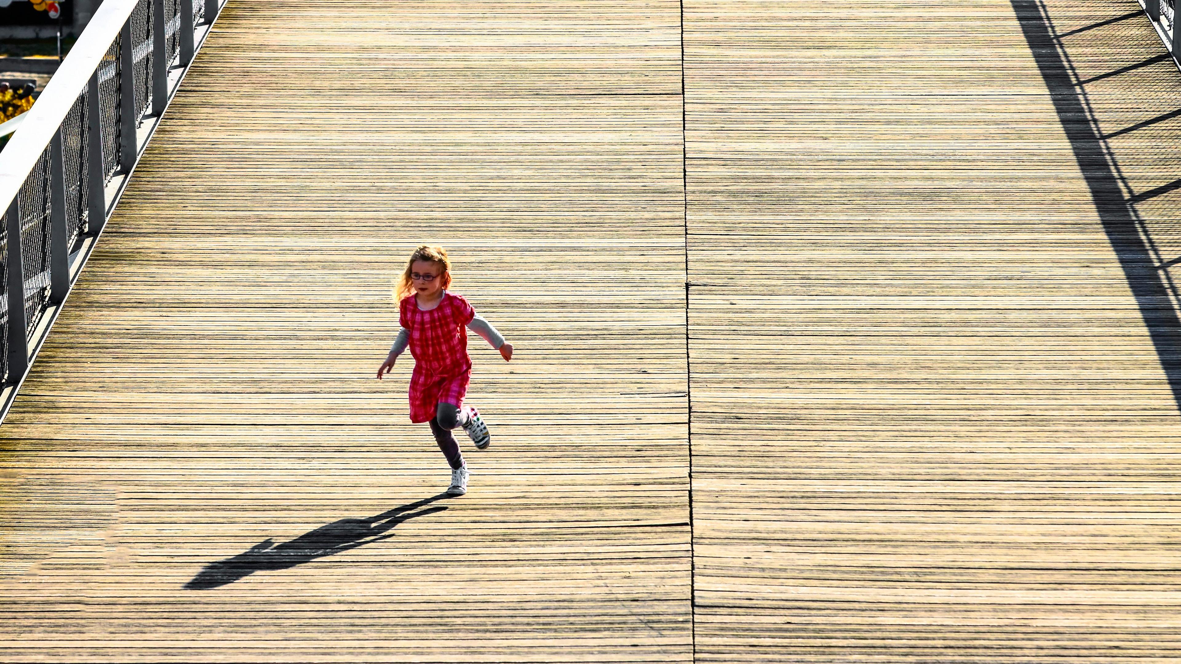A photo of a child in a red dress running on a wooden bridge with adults walking in the background under a clear sky.