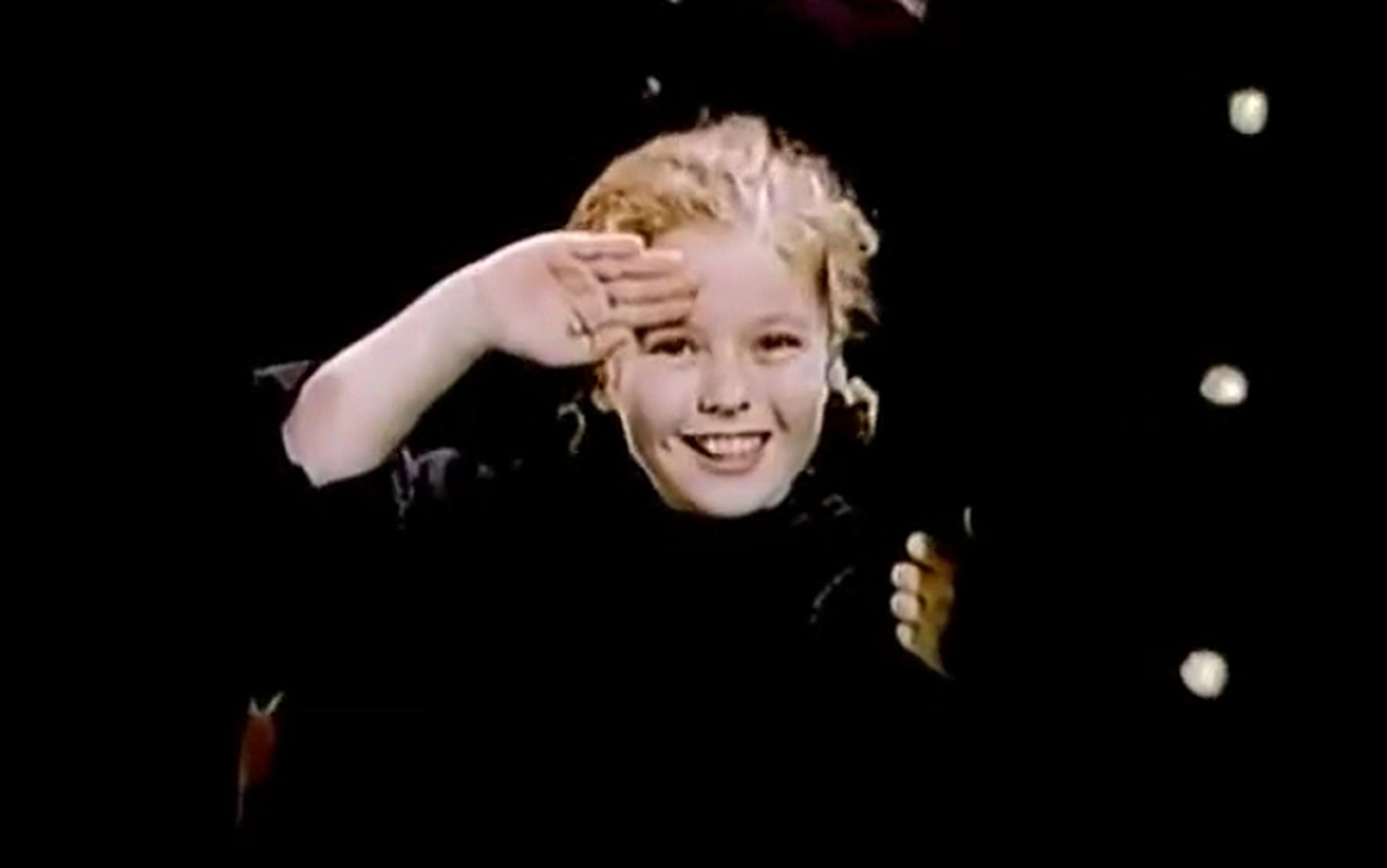 A film still showing a cheerful child with blonde hair smiling saluting. The background is dark with some light spots.
