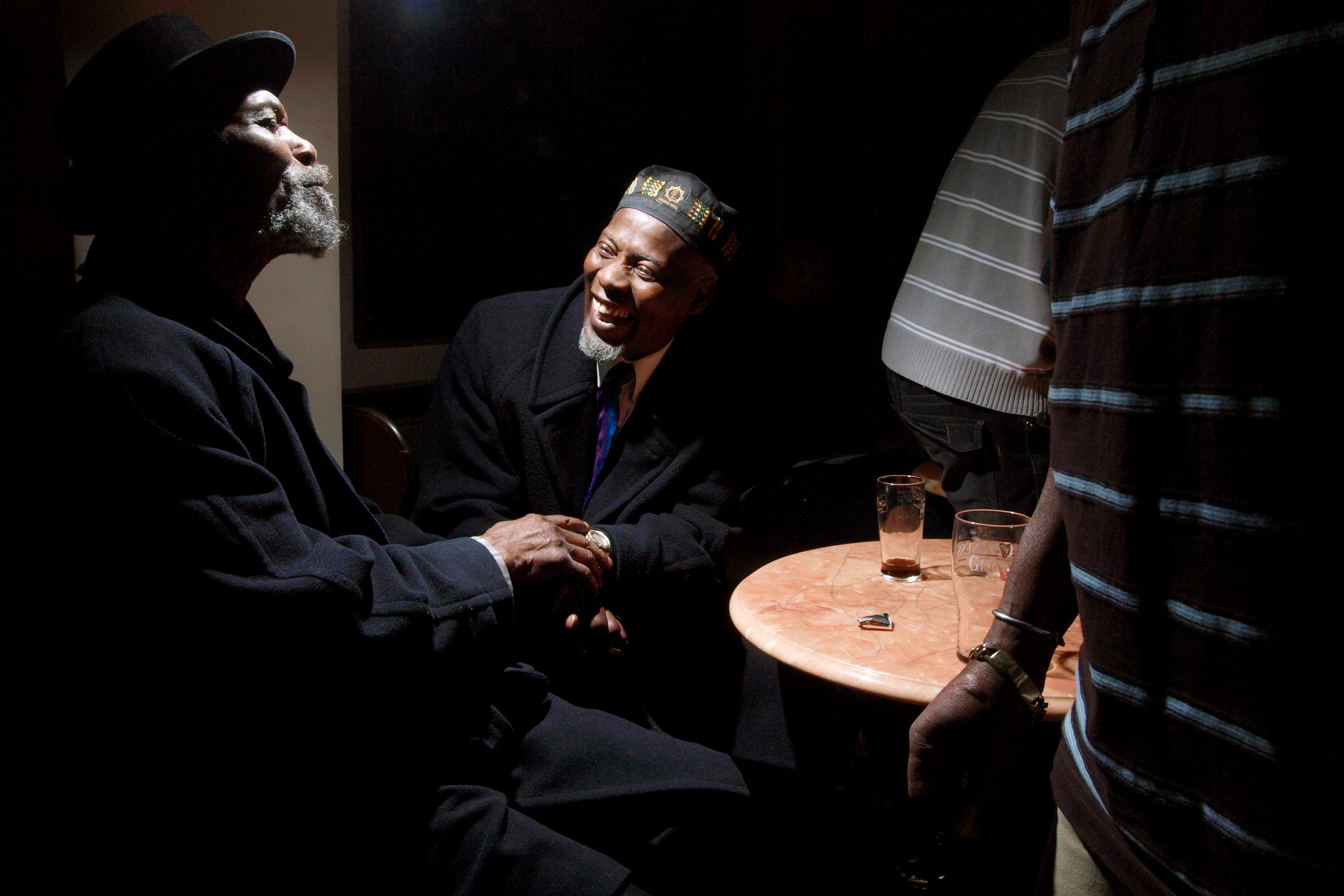 Photo of two men in dark clothing sitting and smiling at a round table in a dimly lit room with glasses.