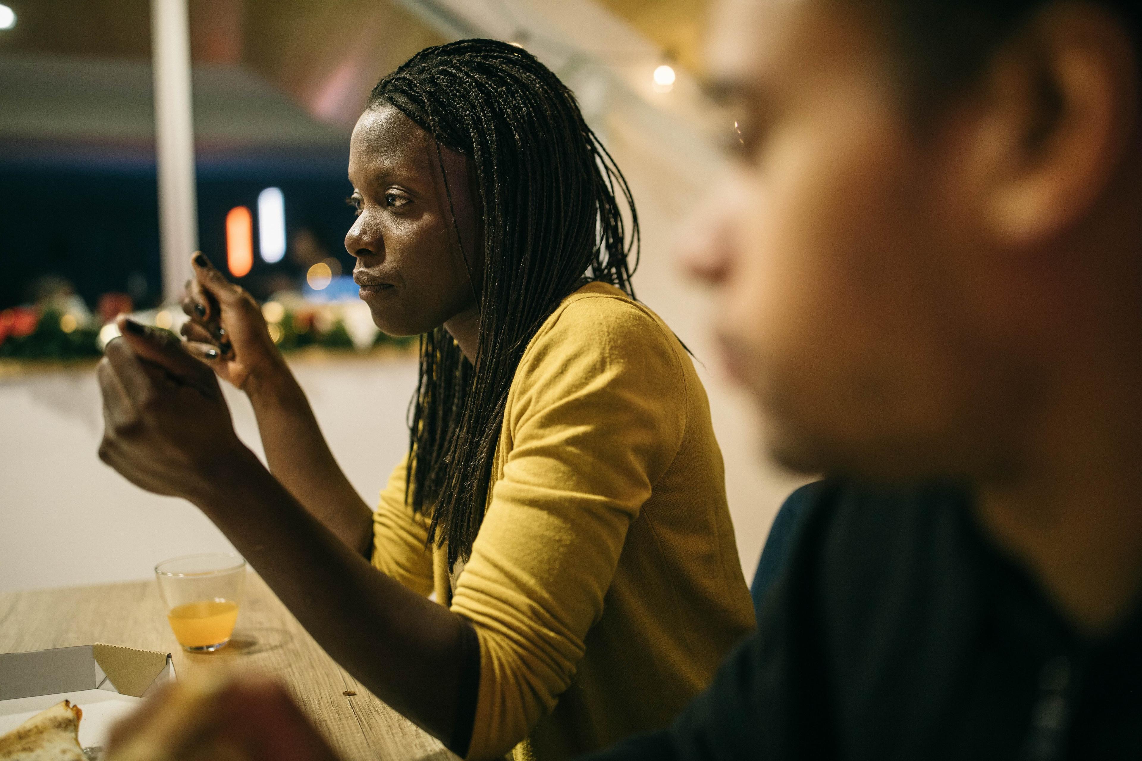 Photo of a woman in a yellow top using her phone at a table, with a blurred person in the foreground.
